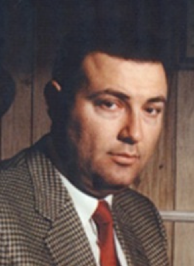 A man with dark hair wearing a checkered jacket and red tie, looking at the camera against a wooden background.