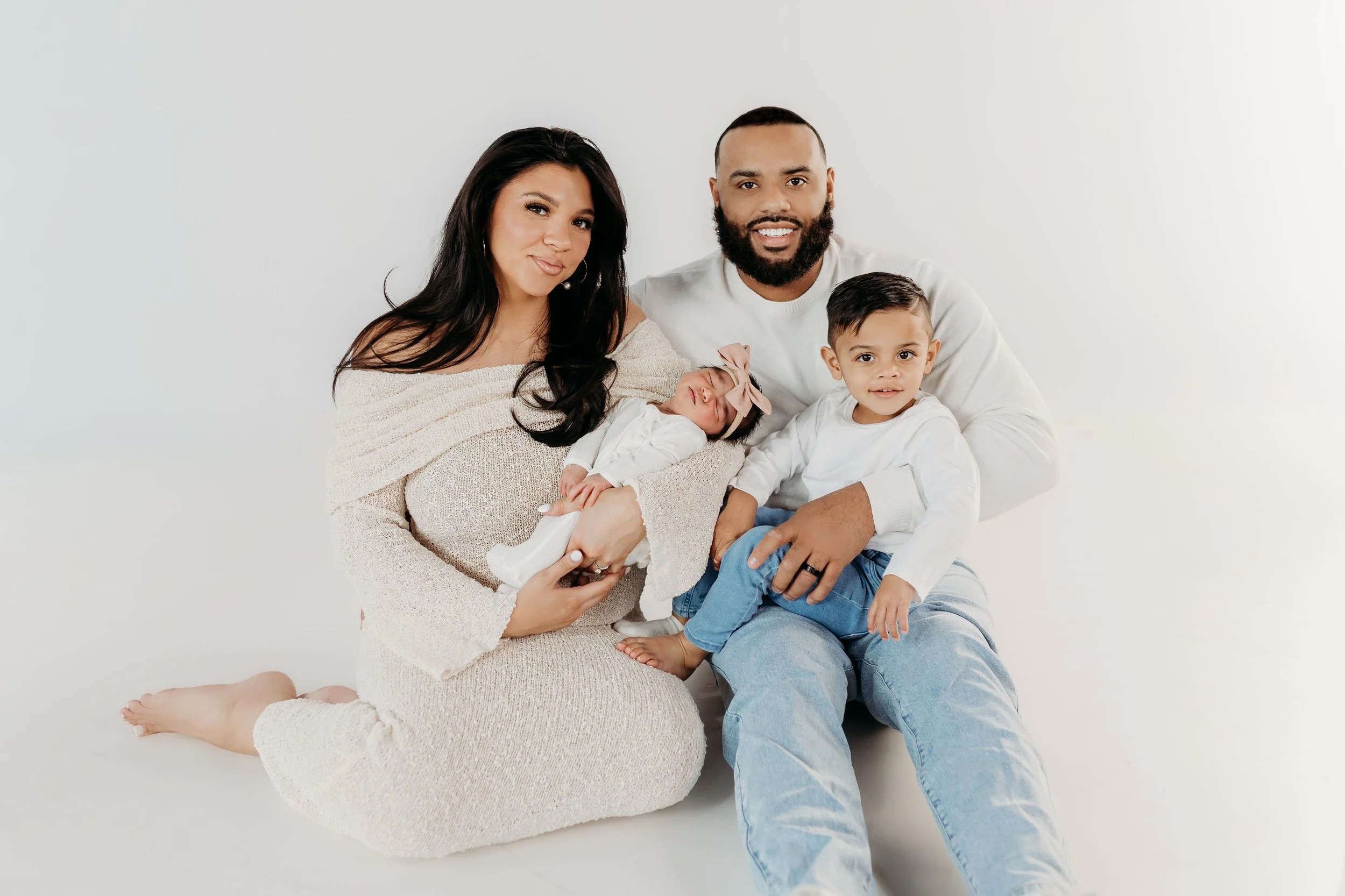 Family portrait of a woman, man, young boy, and newborn baby girl with a pink bow, sitting against a plain white background. The woman is holding the baby, and the young boy is sitting close to the man, all smiling or with relaxed expressions.