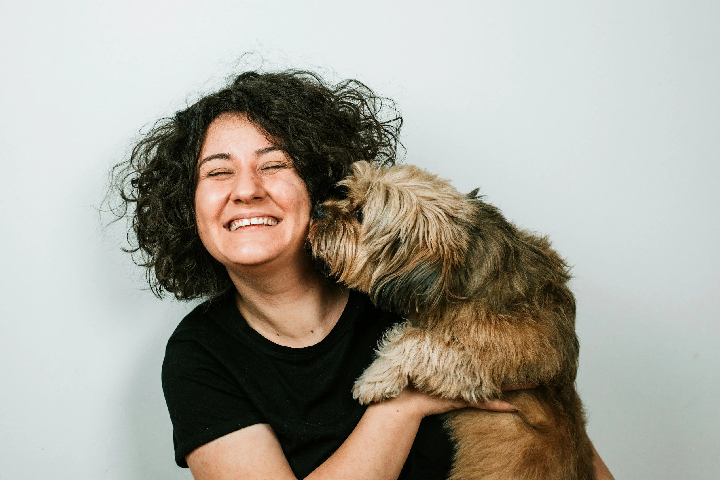 A woman with curly dark hair smiling while holding a large, fluffy brown dog close to her face against a light gray background.