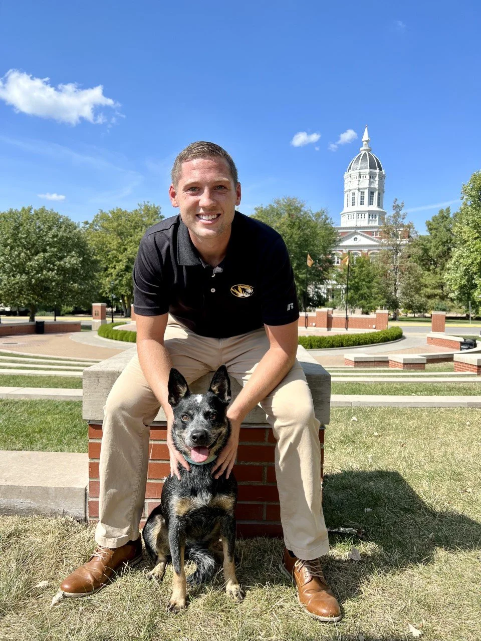 A man in a black polo shirt and beige pants sitting on a brick ledge outdoors, smiling and holding a black and tan dog with pointed ears, against a background of green trees and a white domed building under a blue sky with a few clouds.