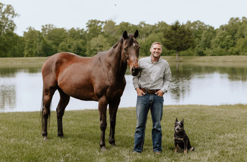 Pet owner standing beside a horse and dog near a quiet lake.