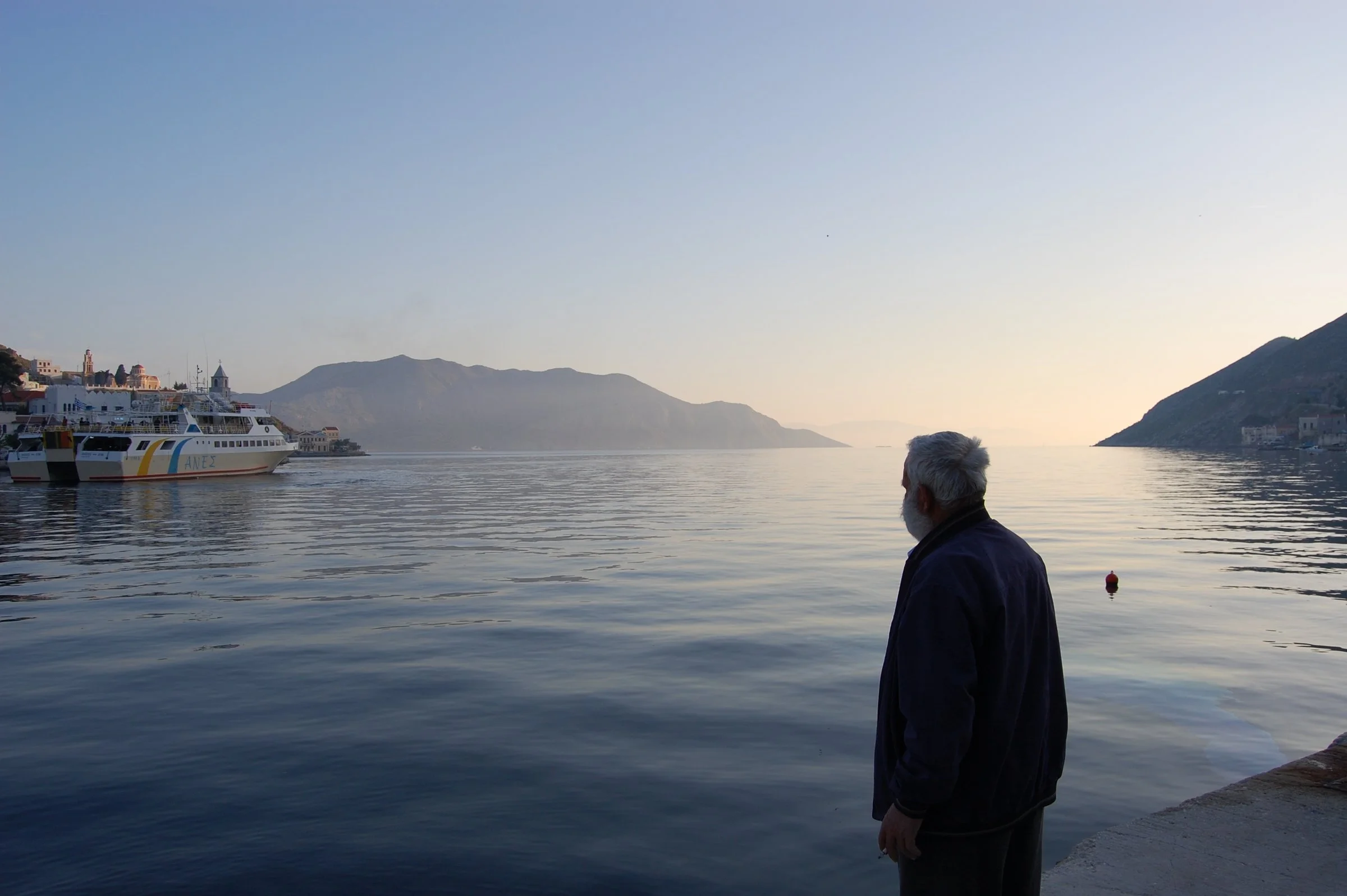 An elderly man standing alone on the harbor of Symi while a ferry sails away across the Aegean Sea.