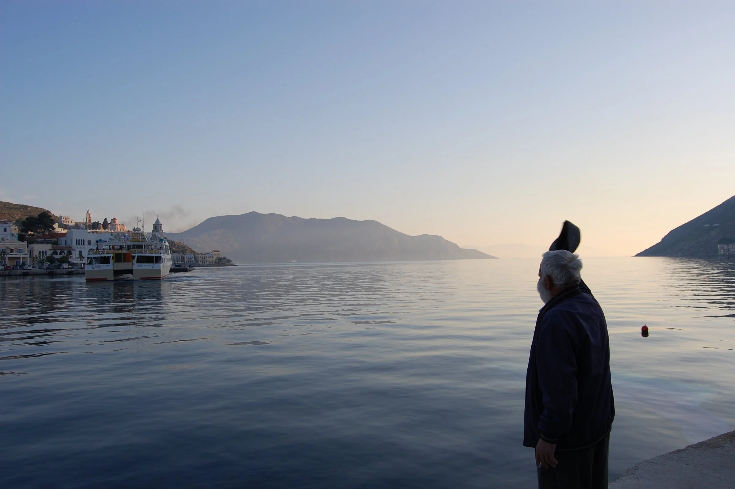 An elderly man watching a departing ferry become smaller on the horizon from the harbor of Symi.