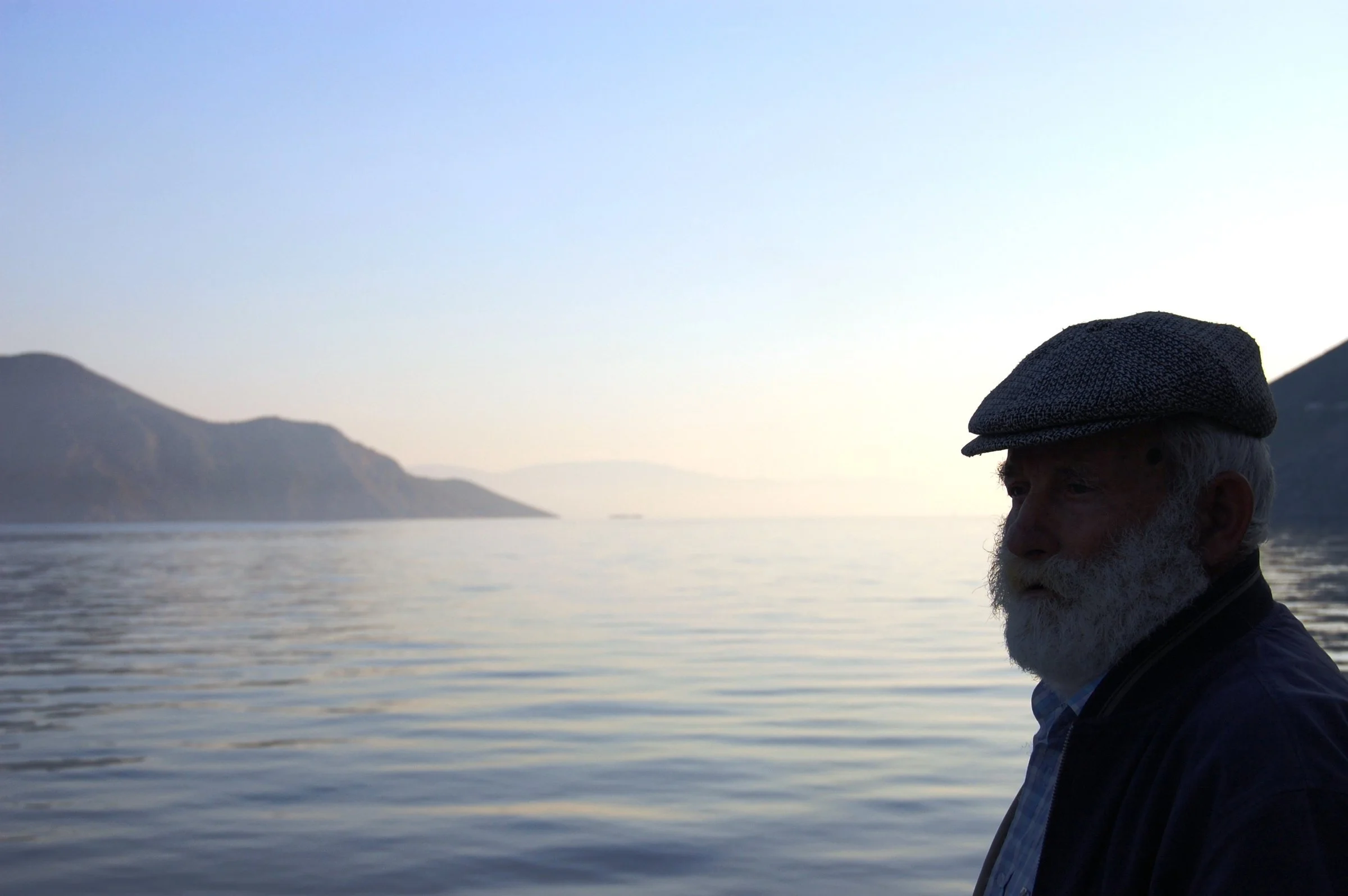 An elderly man standing at the harbor of Symi after a ferry's departure, turning away from the sea toward the island.