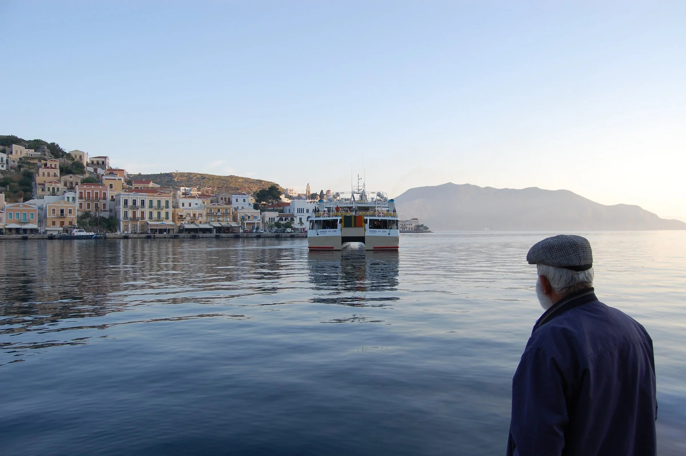 An elderly man standing on the waterfront of Symi as a ferry moves away from the harbor across the calm Aegean Sea.