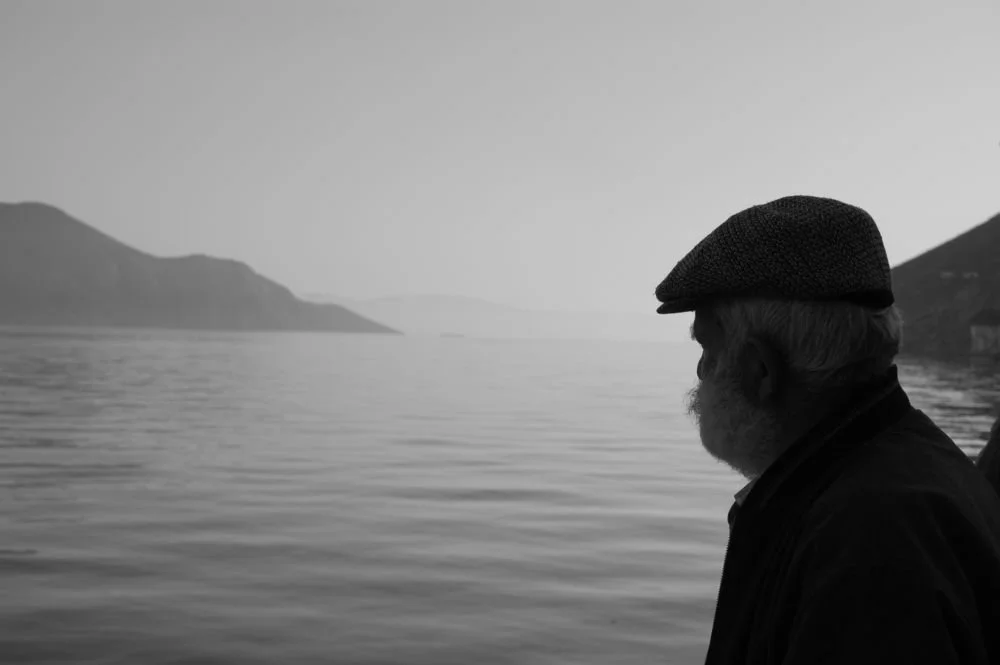 An elderly man looking across the calm waters of Symi Harbor in black and white, moments after a ferry's departure.