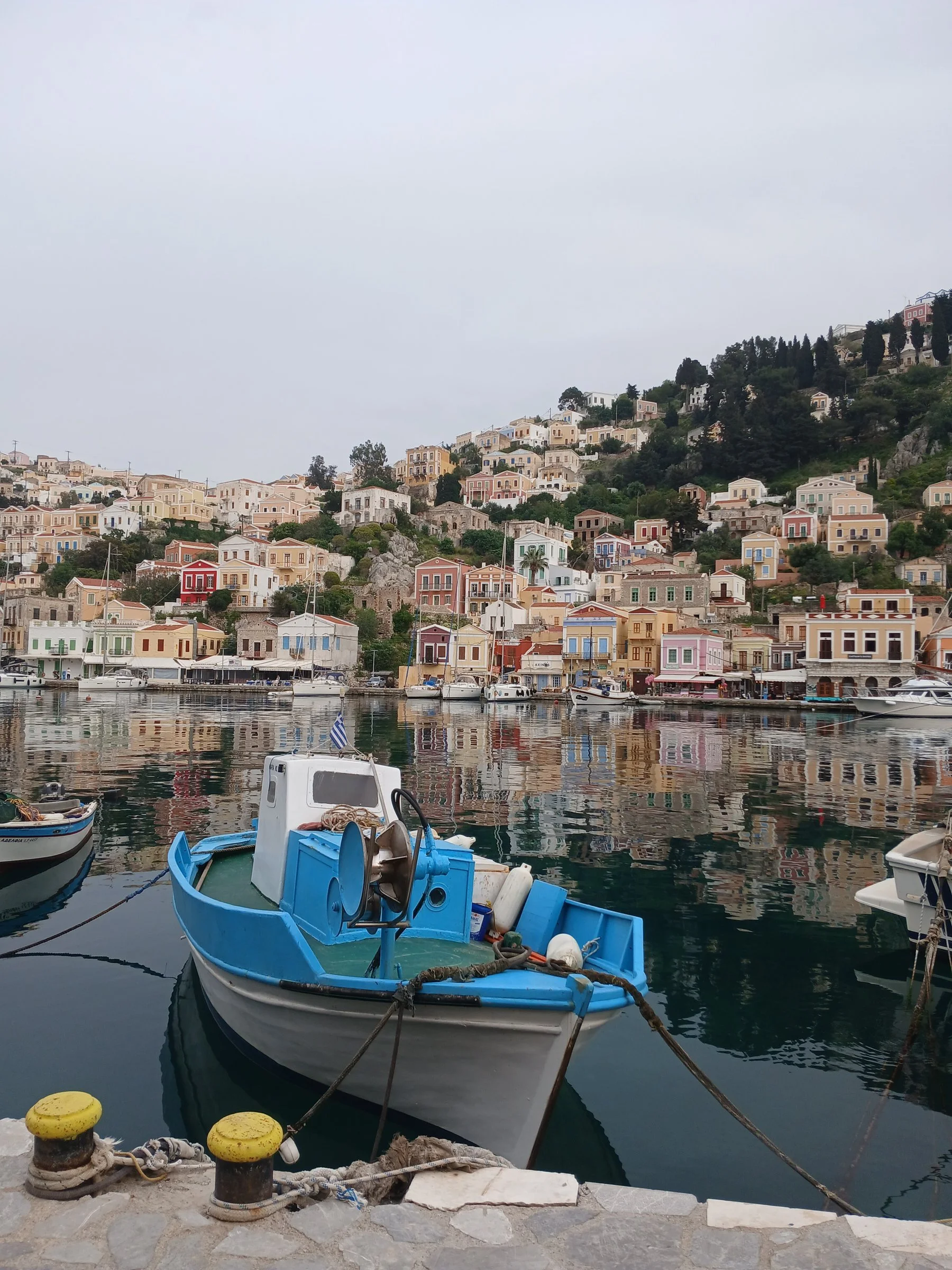 A traditional, salt-worn blue wooden boat gently floating in a quiet island harbor with old stone houses in the background.