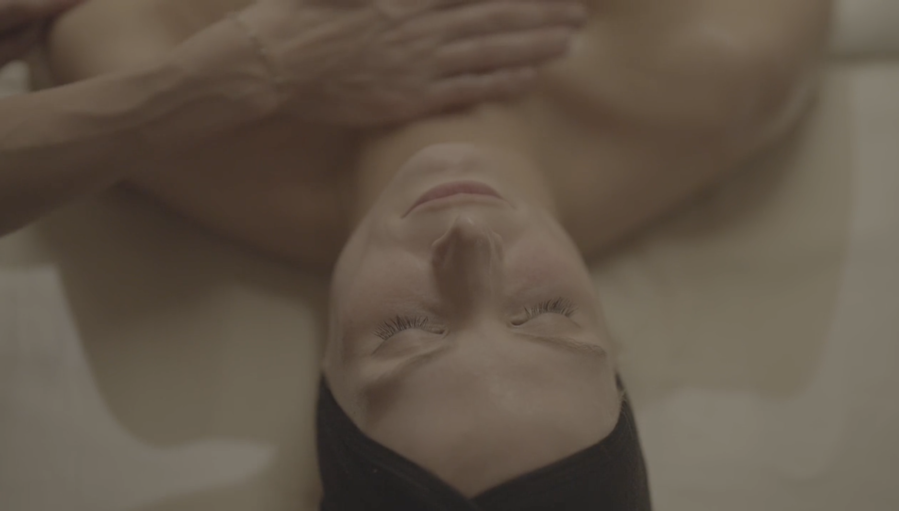 A woman lies on her back with her eyes closed, her hands resting on her chest, and her head tilted slightly to the side.