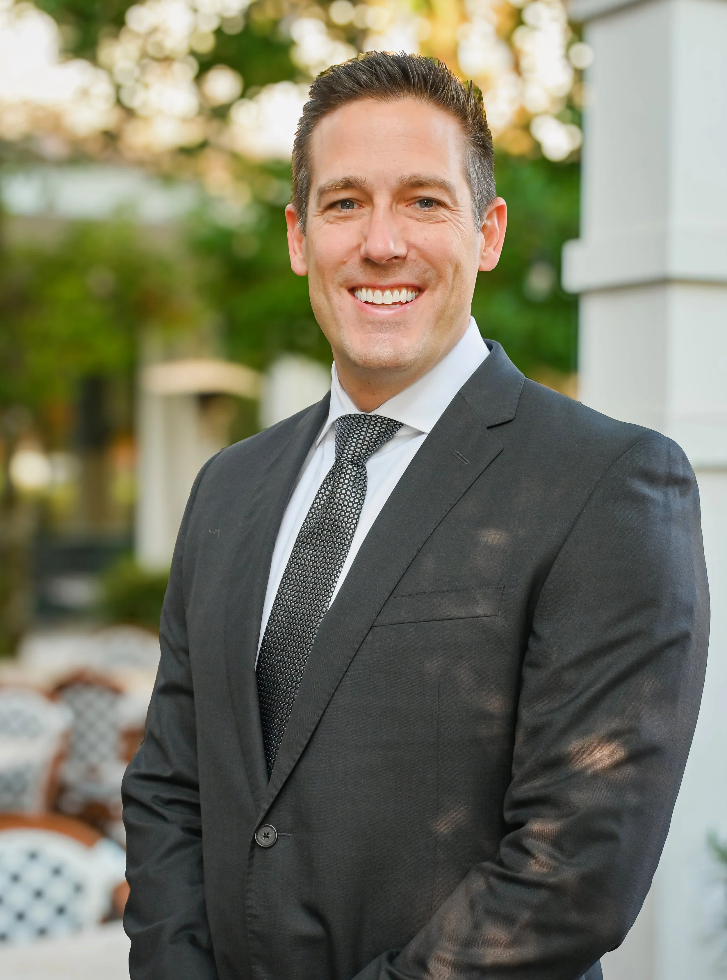 A smiling man in a dark suit with a tie, standing outdoors with a blurred background of trees and house siding.