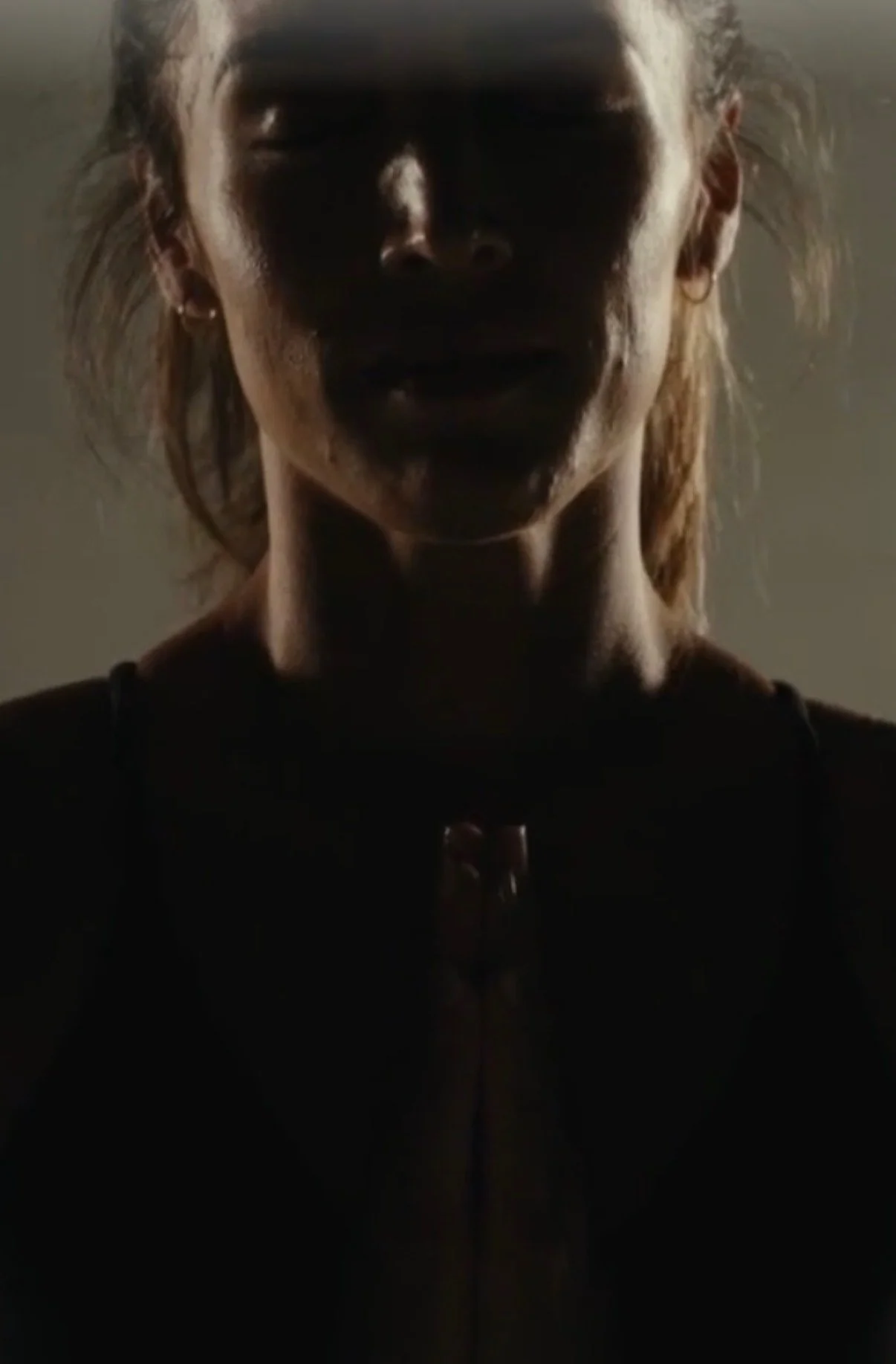 Person with light-colored hair and earrings, wearing dark clothing, in low lighting, with shadows covering part of their face.