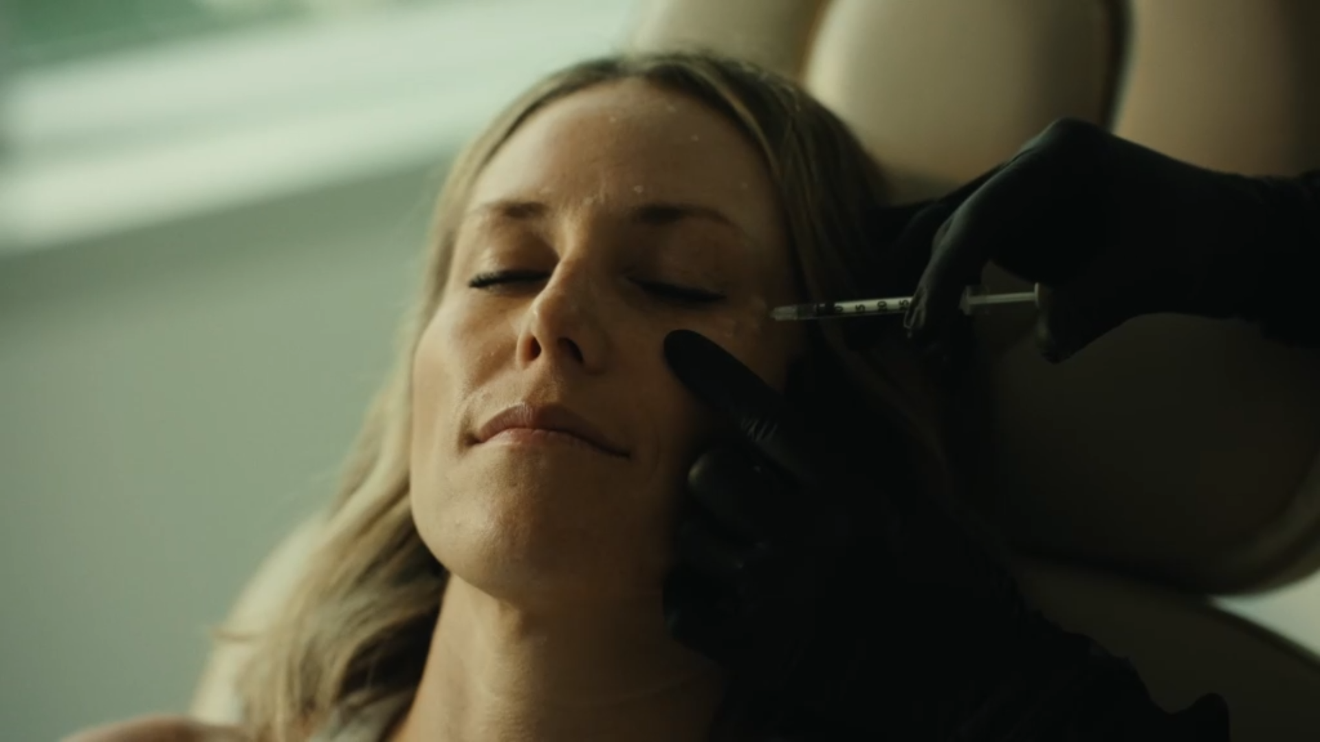 A woman with closed eyes receiving a cosmetic injection in her face from a person wearing black gloves.