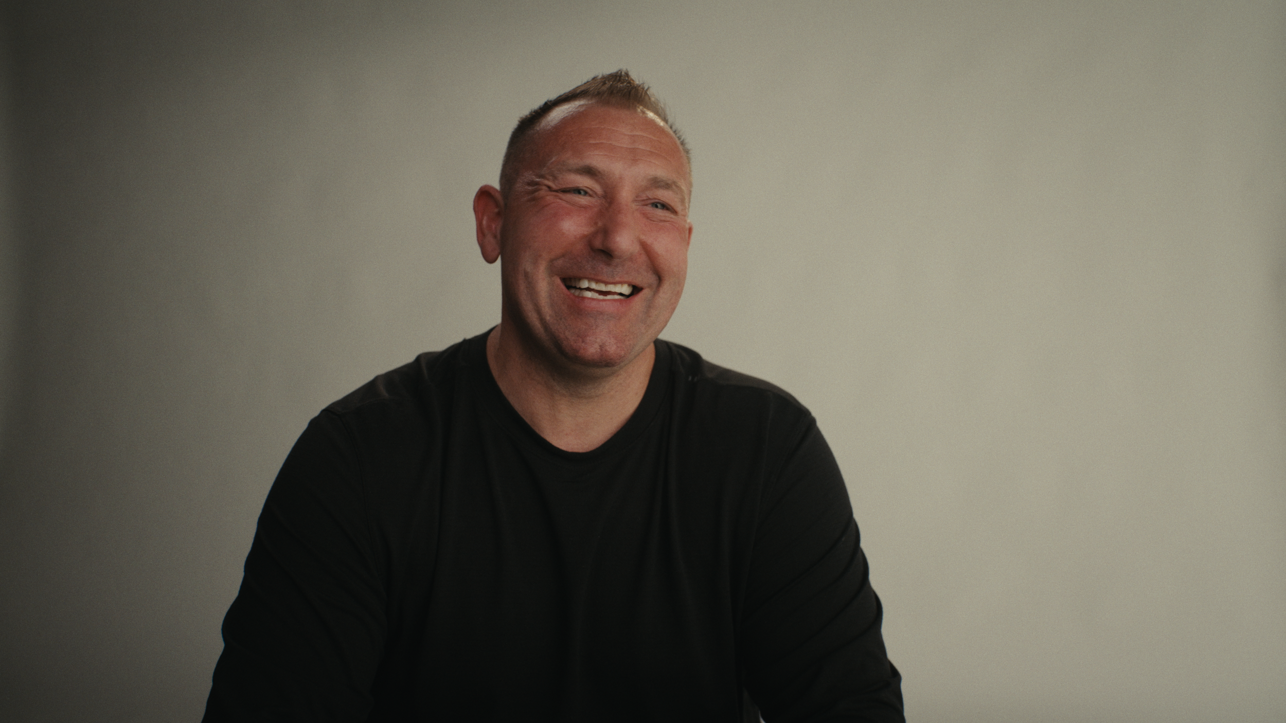 A man with short hair and a big smile, wearing a black shirt, sitting against a plain light-colored wall.