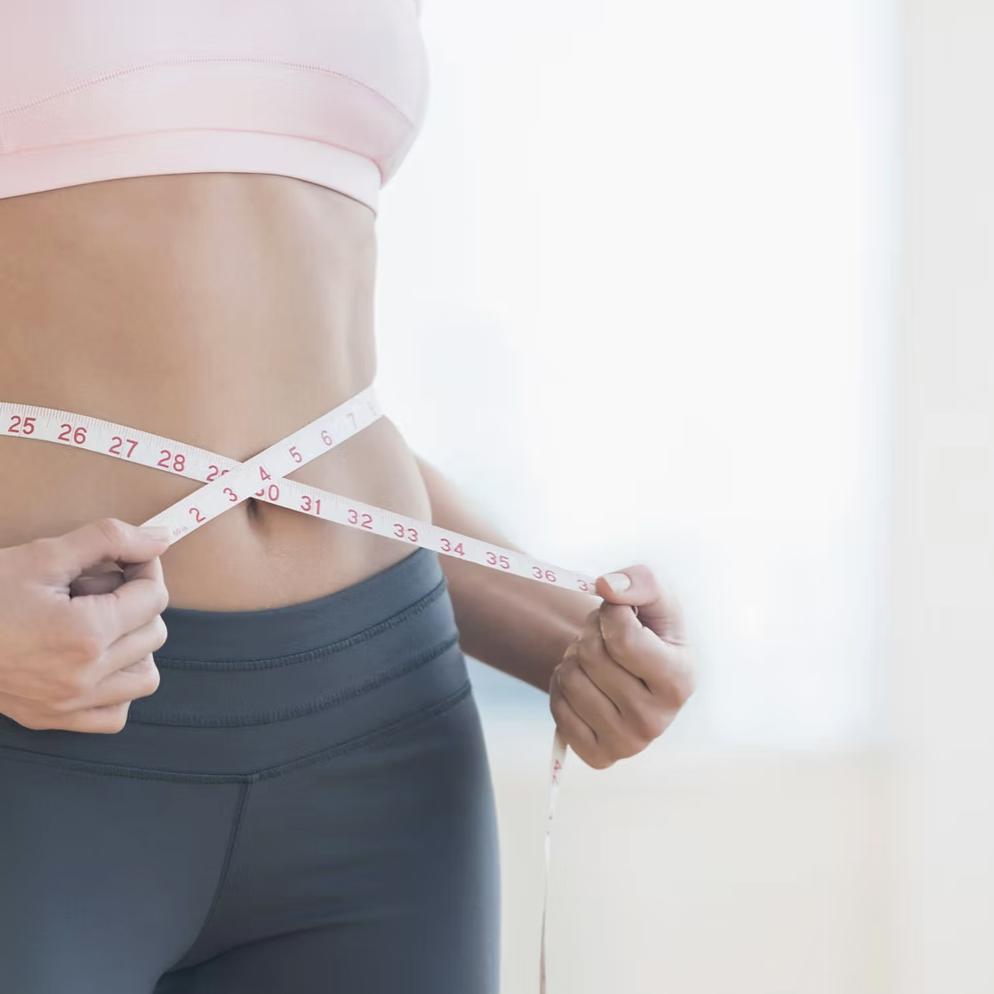 A woman measuring her waist with a white measuring tape while wearing a pink sports bra and dark athletic leggings.