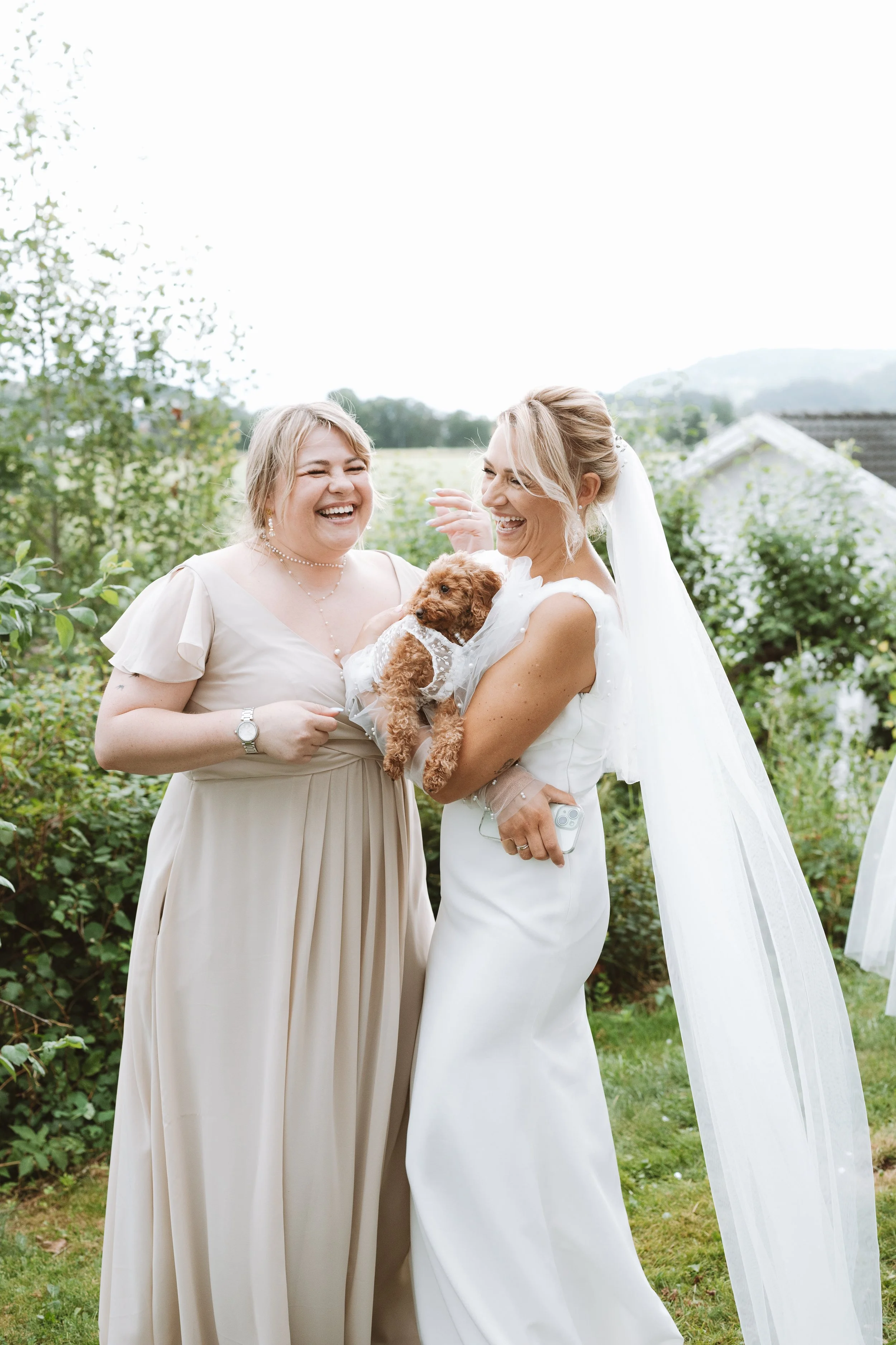 Two women, one in a wedding dress and the other in a beige dress, are smiling and holding a small brown puppy in an outdoor garden setting.