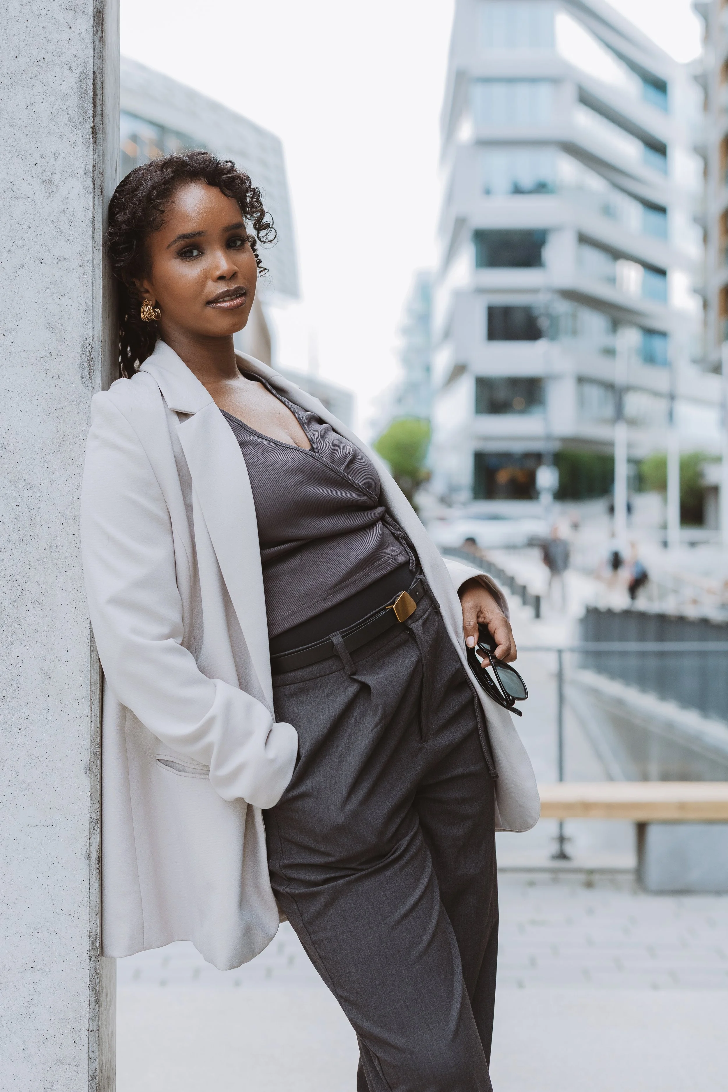 A woman with curly hair and gold hoop earrings leaning against a concrete wall in an urban setting with modern white buildings in the background. She is wearing a white blazer over a dark top and gray pants, holding sunglasses in her right hand.