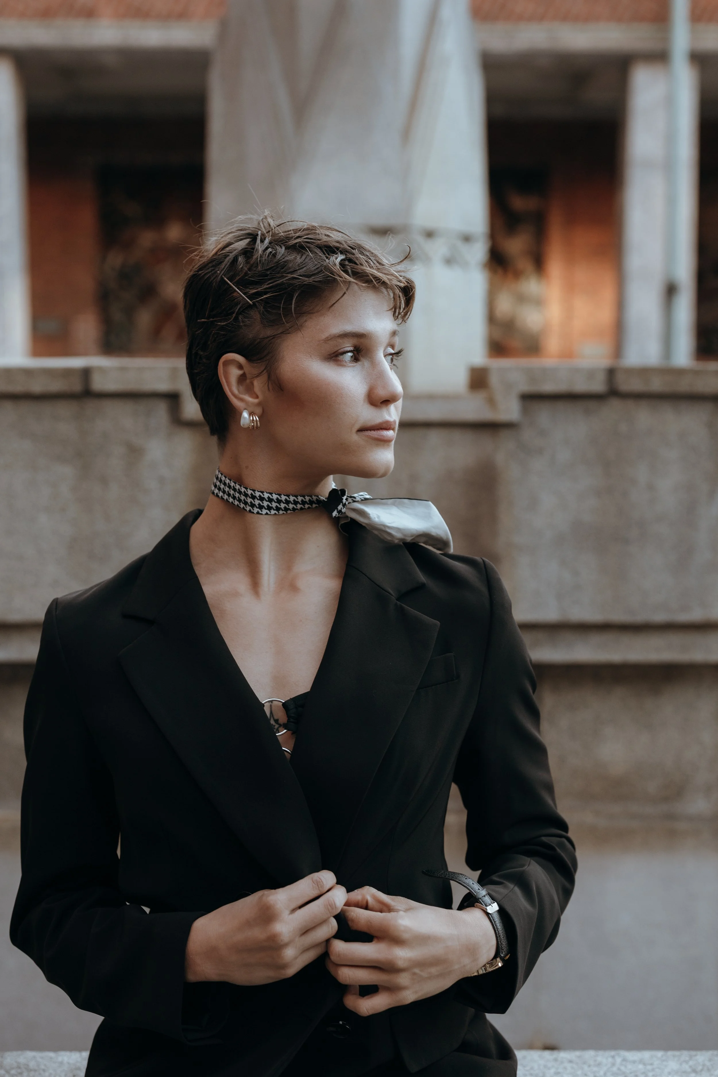 A woman with short brown hair, wearing a black blazer, patterned choker, earrings, and a watch, standing outdoors in front of a brick building with concrete structures.