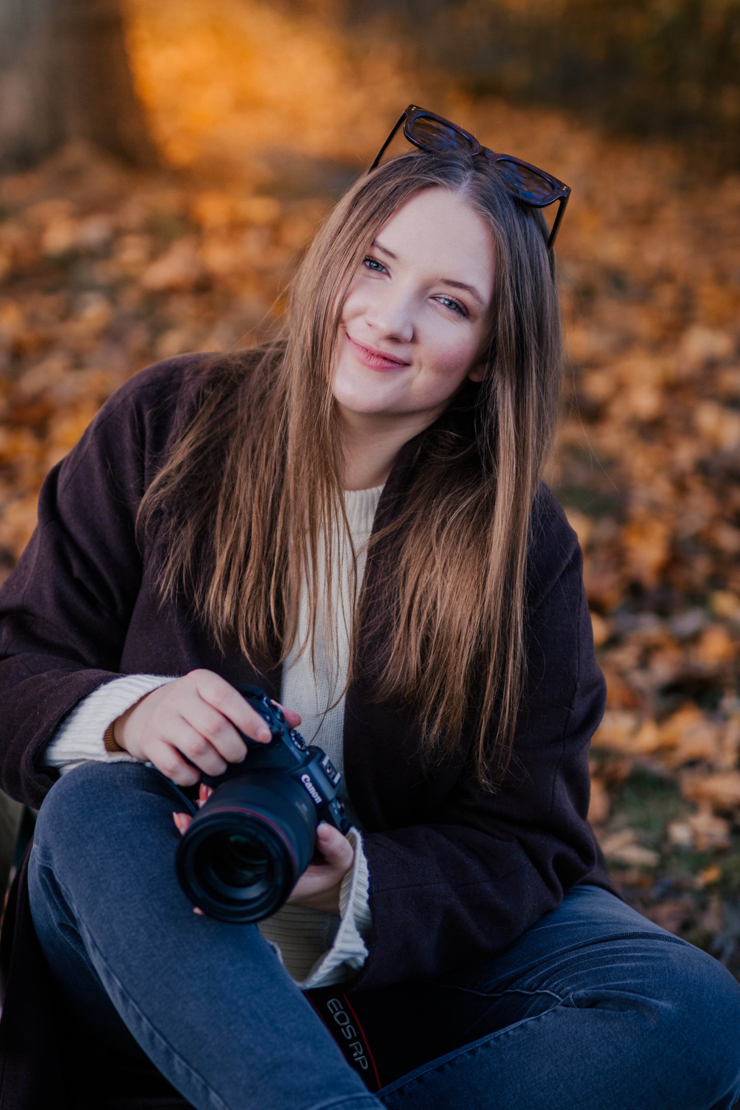 A young woman with long brown hair and blue eyes smiling outdoors in park in Oslo during autumn, sitting on the ground with fallen leaves, holding a professional camera in her hand, wearing glasses on her head, a cream-colored sweater.