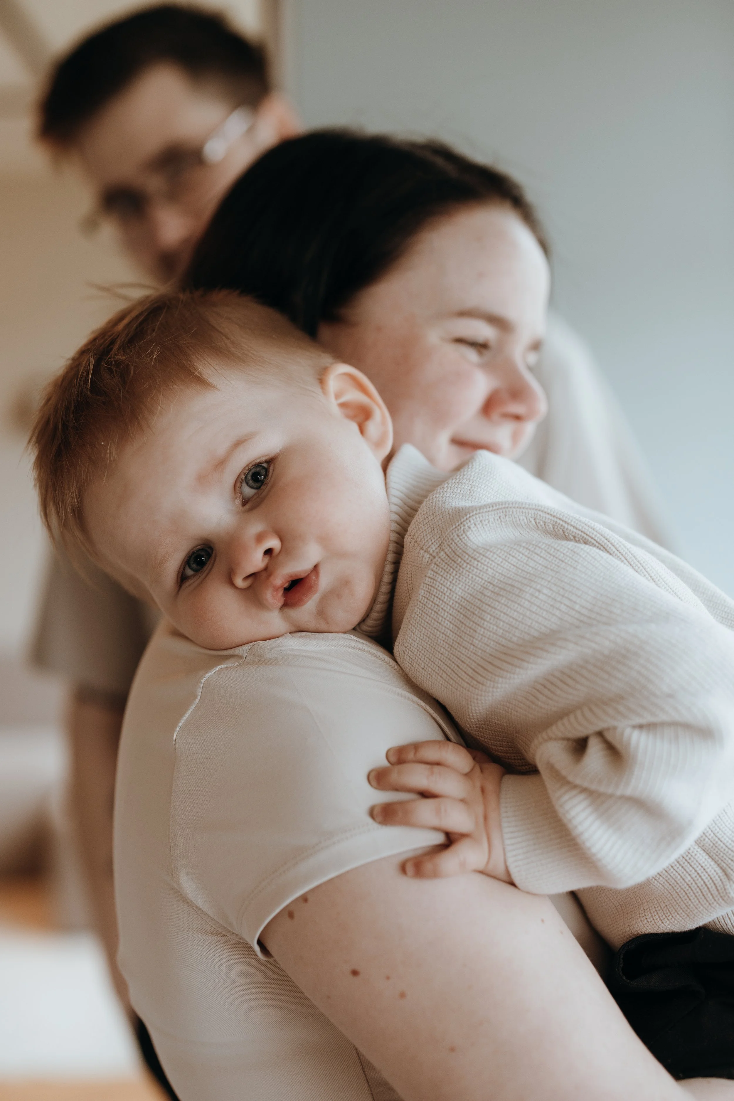 Close-up of a woman holding a young child with blonde hair and blue eyes, smiling and leaning against her chest, in a cozy indoor setting.