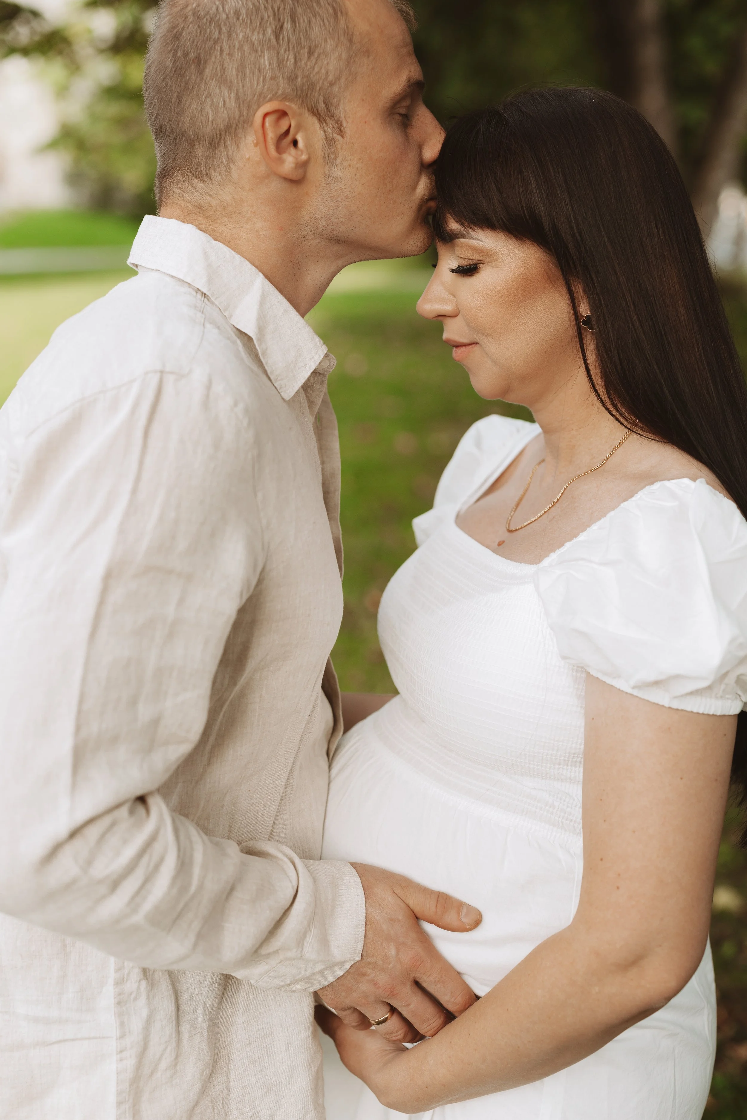 A couple standing outdoors, with the man kissing the woman's forehead as they hold hands and gaze down at her pregnant belly.