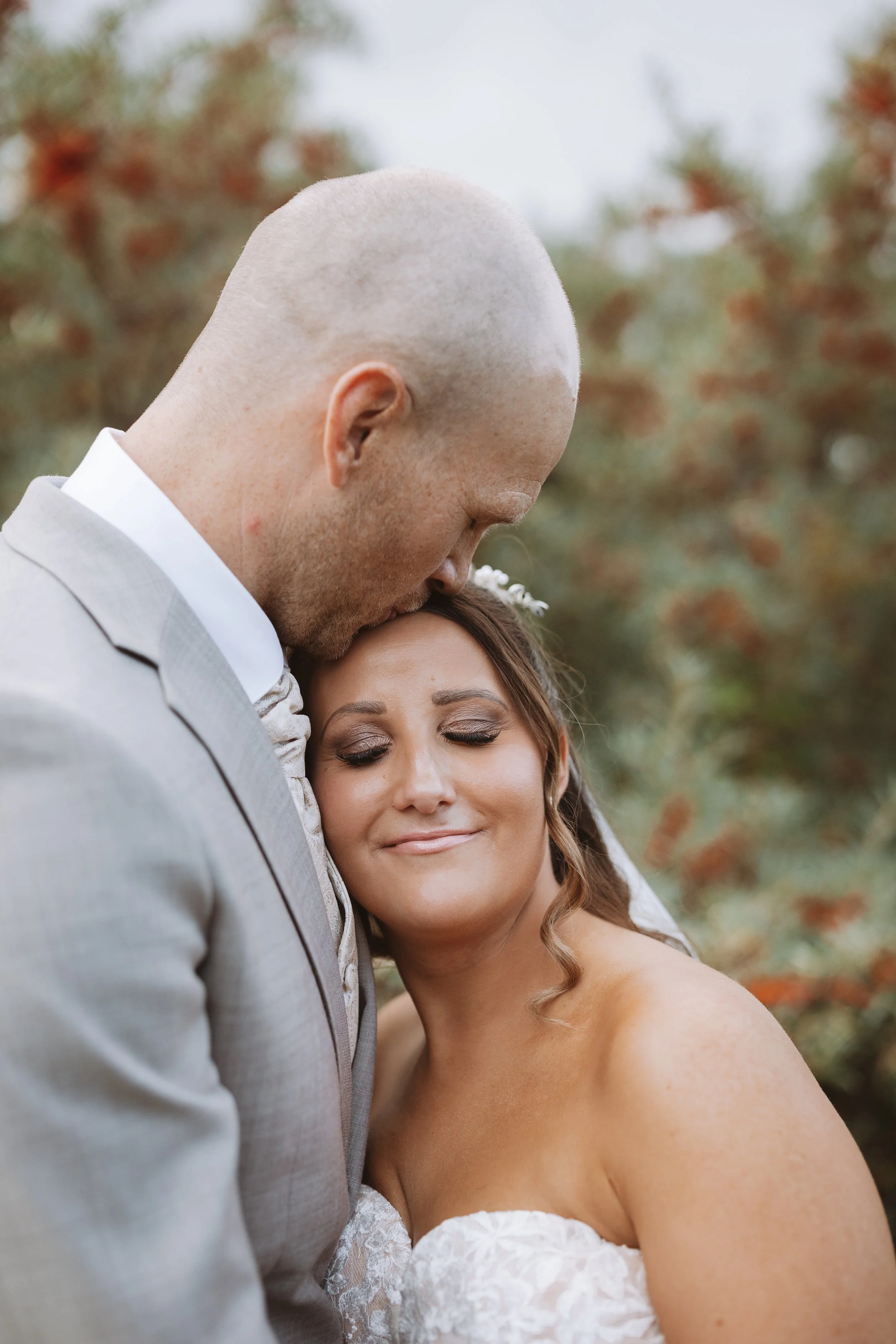 A close-up of a couple during a wedding, with the man kissing the woman's forehead and the woman smiling softly with her eyes closed, outdoors with blurred autumn leaves in the background.