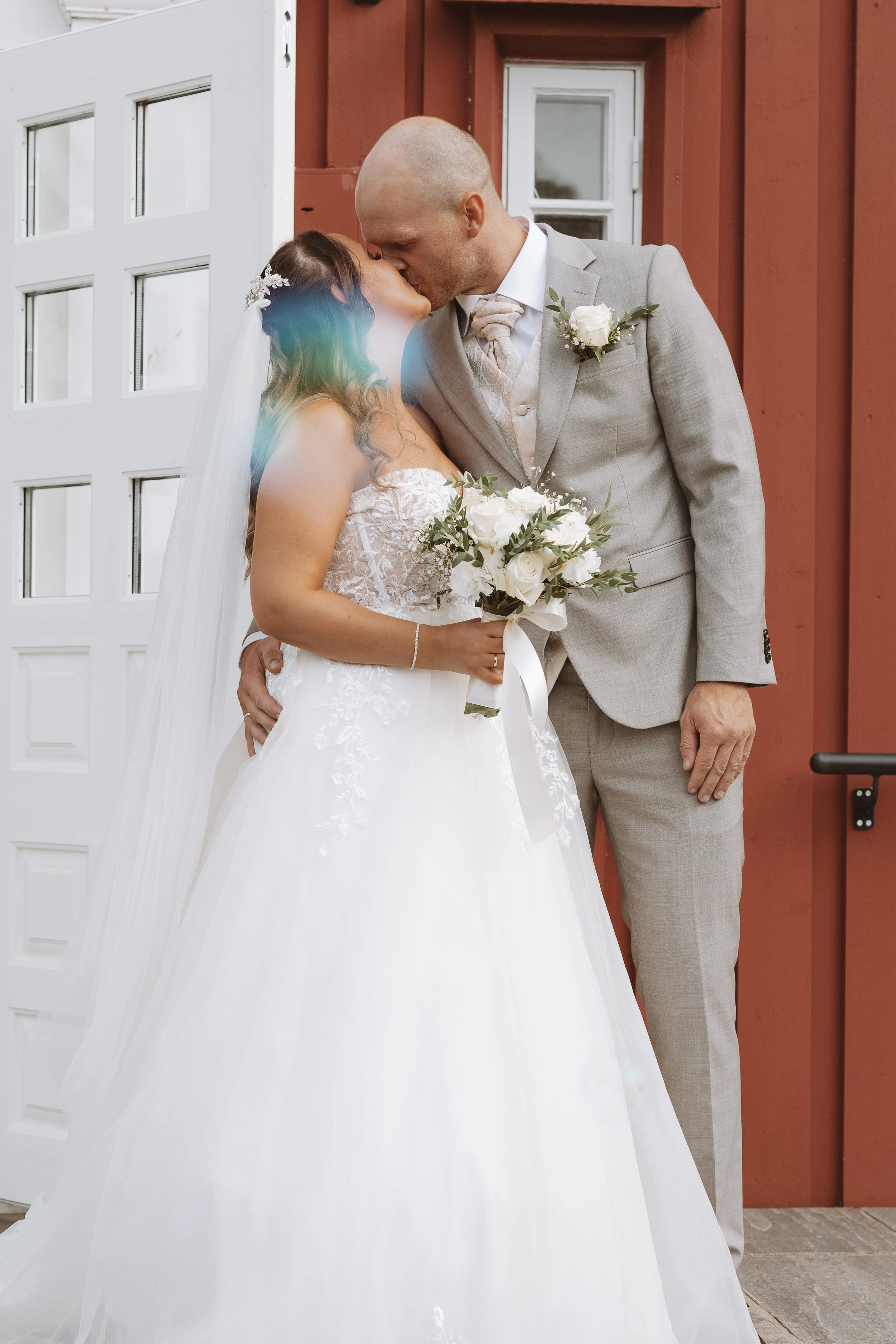 A bride and groom share a kiss outside a building with a red wooden wall and white door.