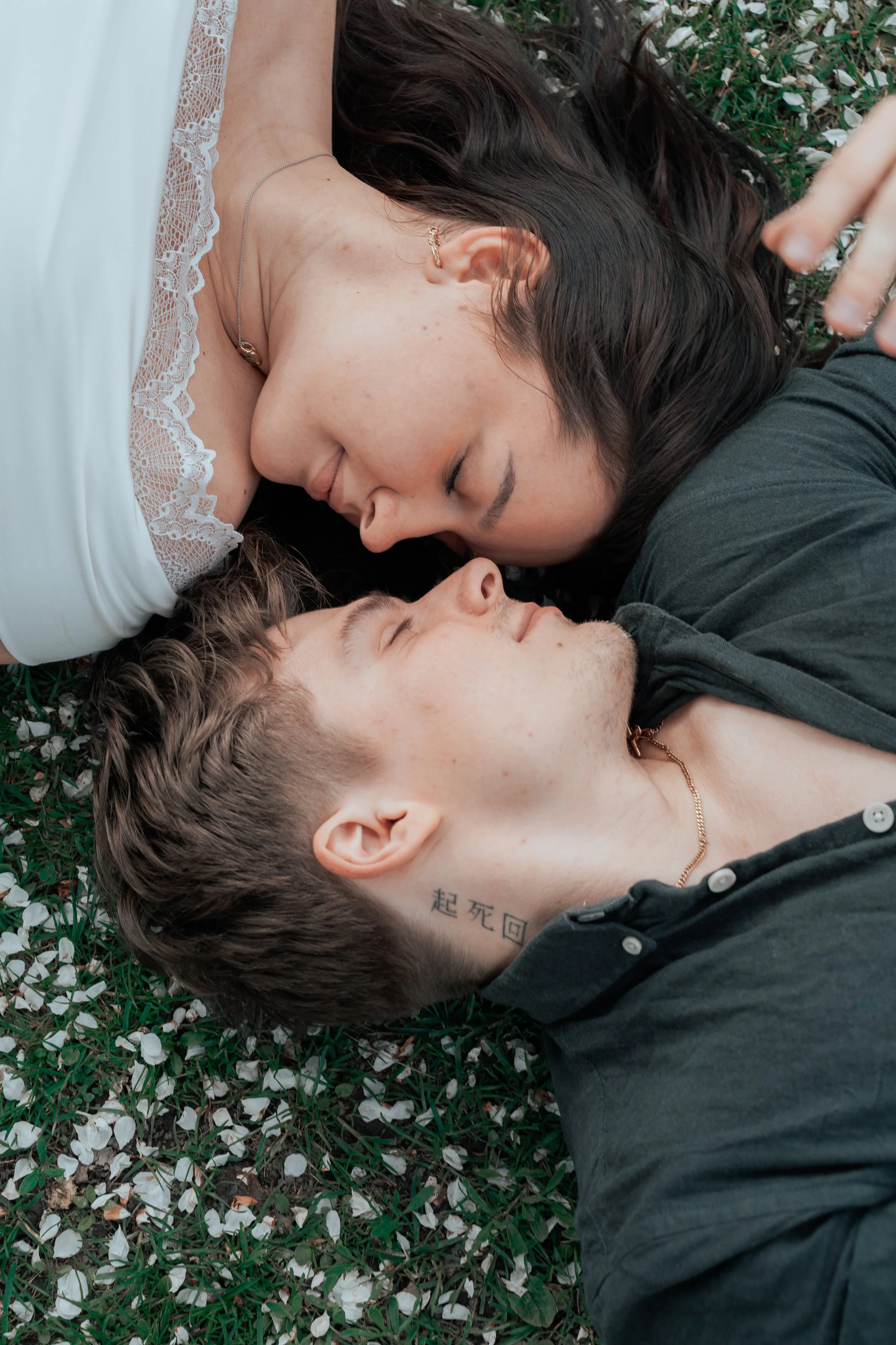 A woman and a man lying face to face on the grass with white flowers, nose to nose, with their eyes closed, appearing intimate and peaceful.