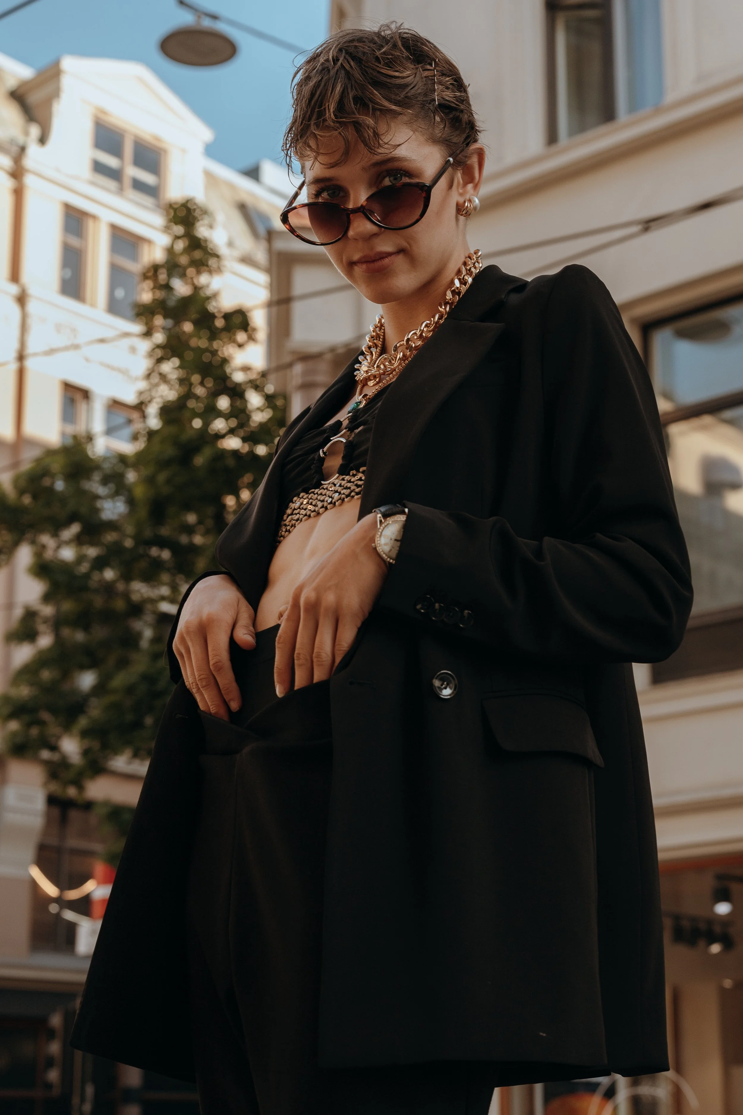A woman with short wet hair wearing black sunglasses, a black blazer, gold jewelry, and a watch, posing outdoors in front of a city building during daytime.