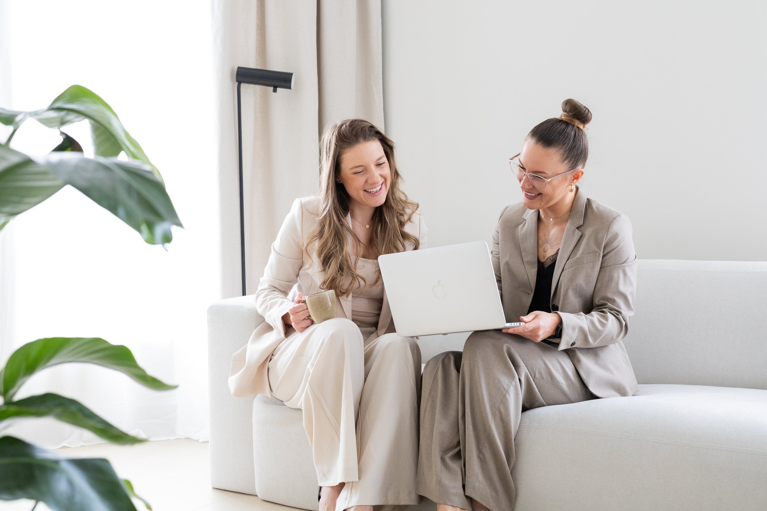 Two women sitting on a white couch in a bright, modern room, sharing a moment of laughter while looking at a laptop. One woman holds a mug, and there are large green plants in the foreground.