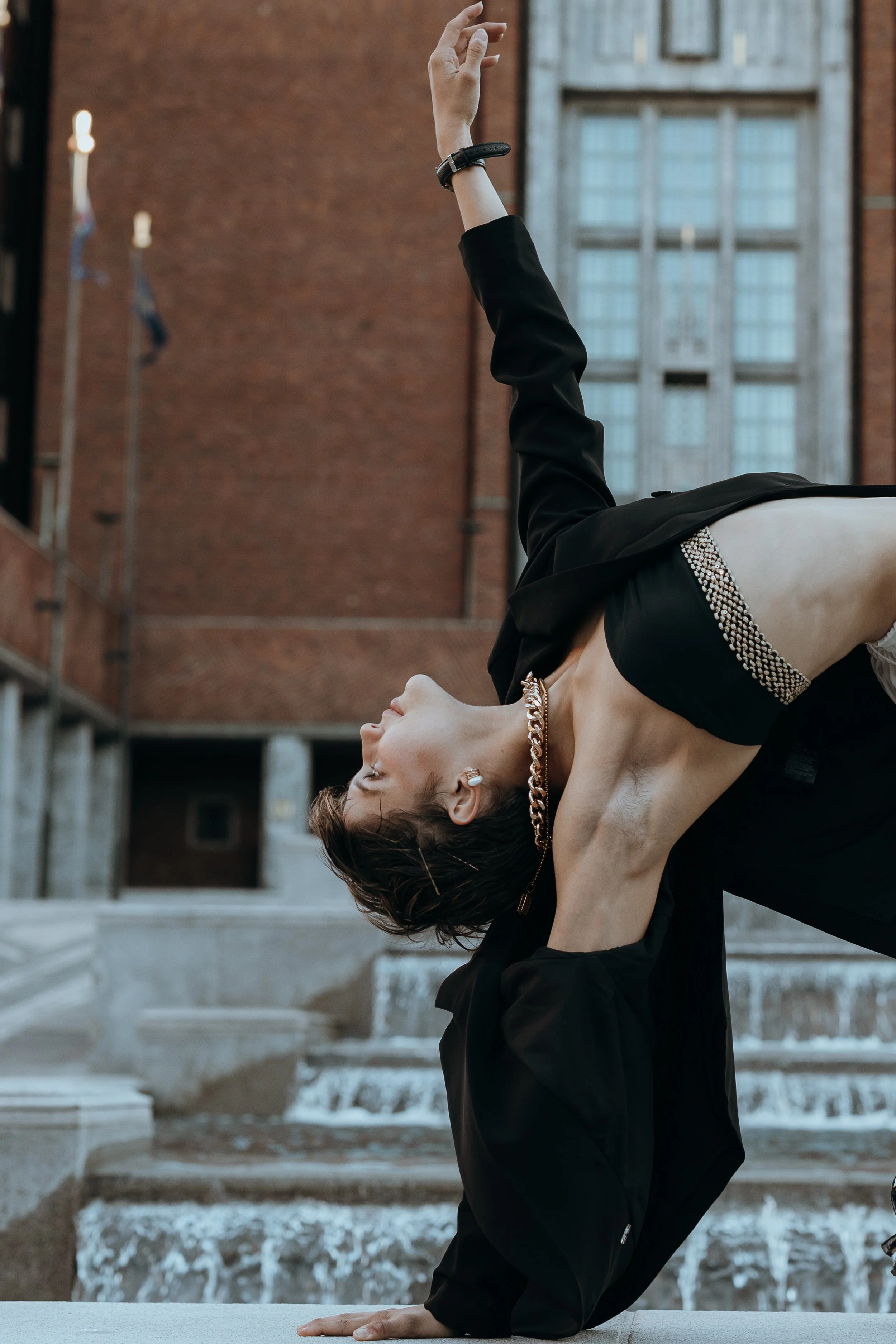 A person practicing yoga outdoors on steps with water fountain, brick building, and large window in the background, doing a backbend pose with one arm and head touching the ground.