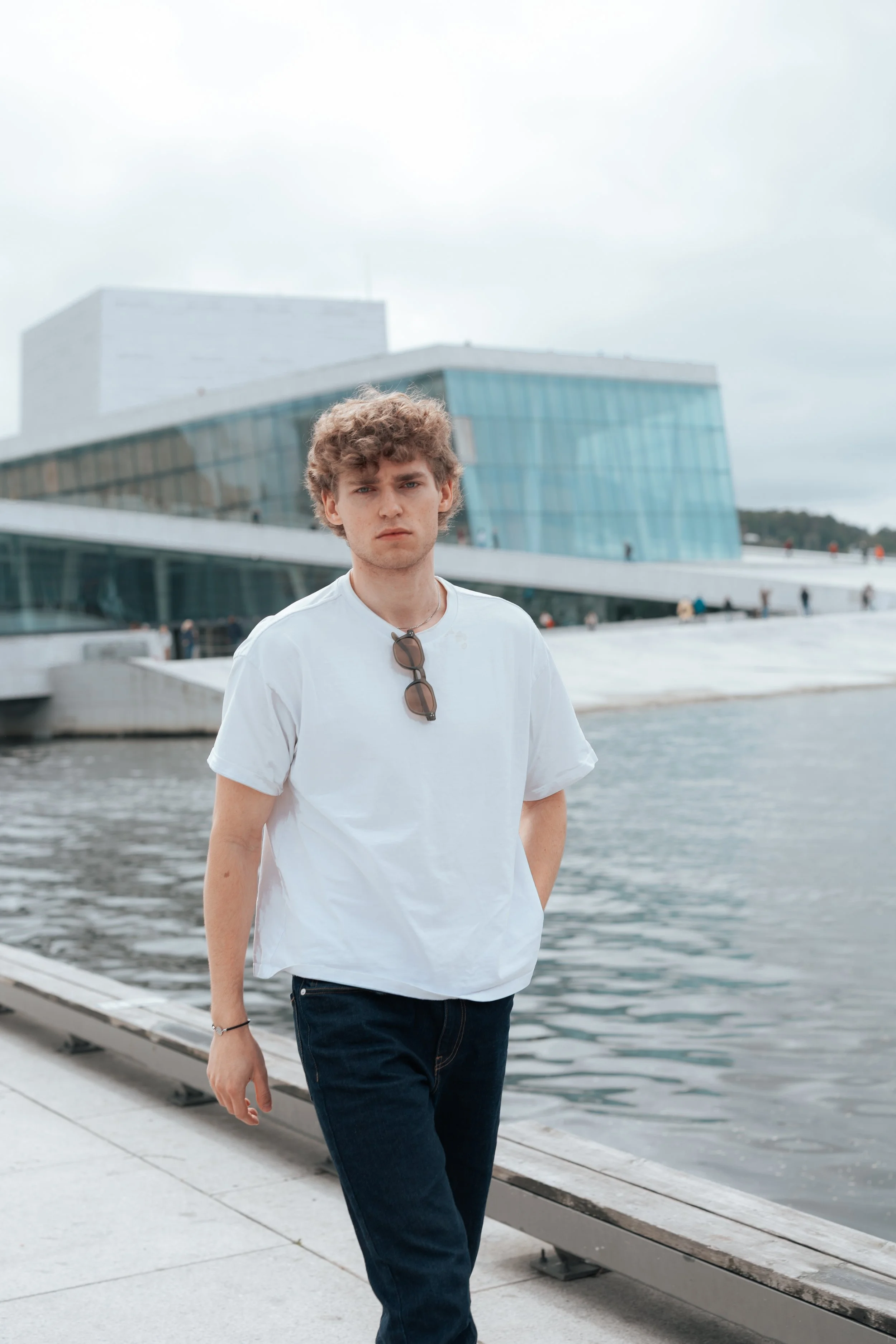A young man with curly hair, wearing a white t-shirt with sunglasses hanging on his collar, stands by a body of water with modern glass buildings in the background.