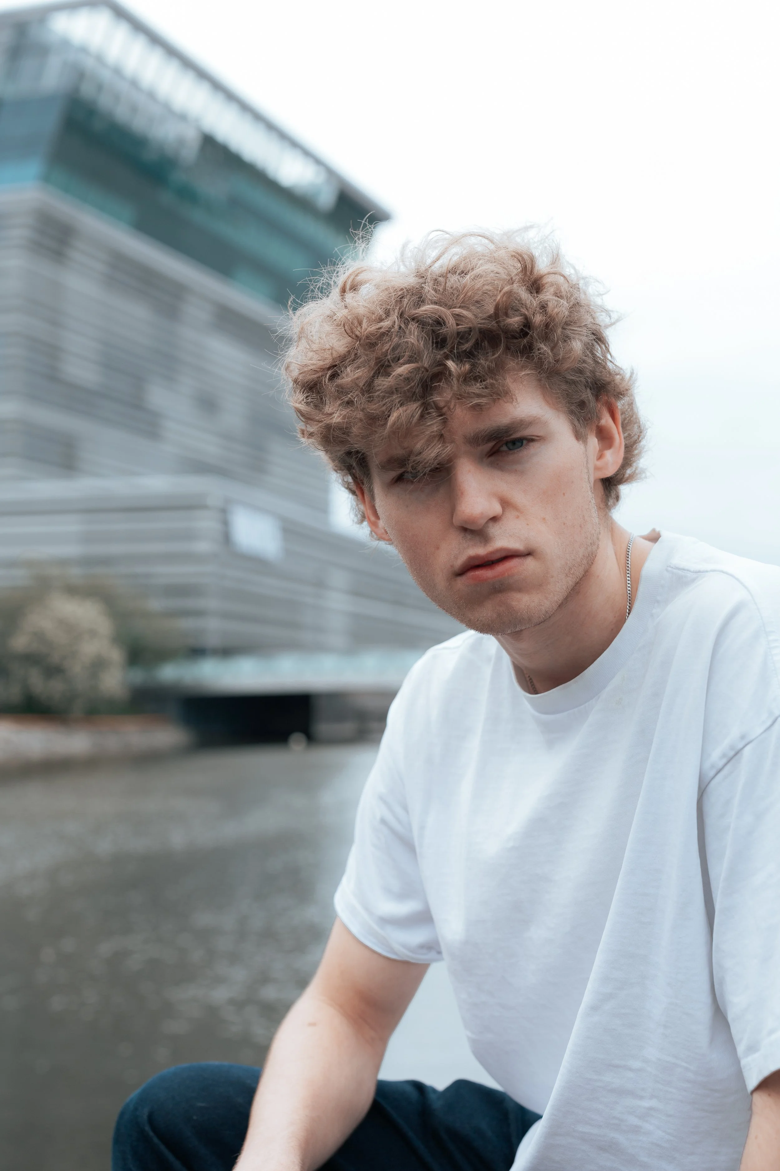 Portrait of a young man with curly light brown hair wearing a white t-shirt, sitting outdoors near a modern building of Munch musem in Oslo.