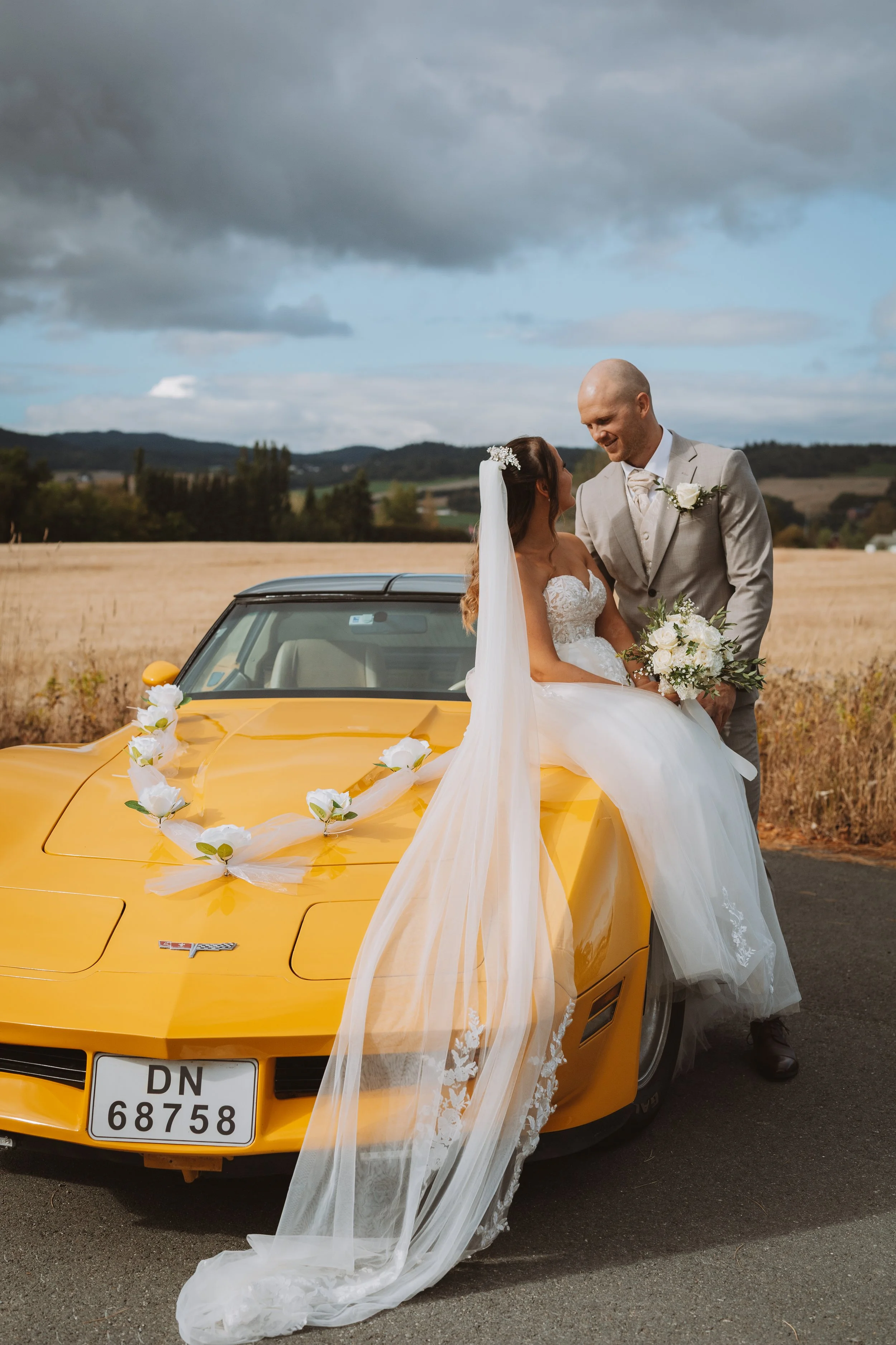 A bride and groom standing next to a yellow sports car decorated with white flowers in a rural field under gray clouds.