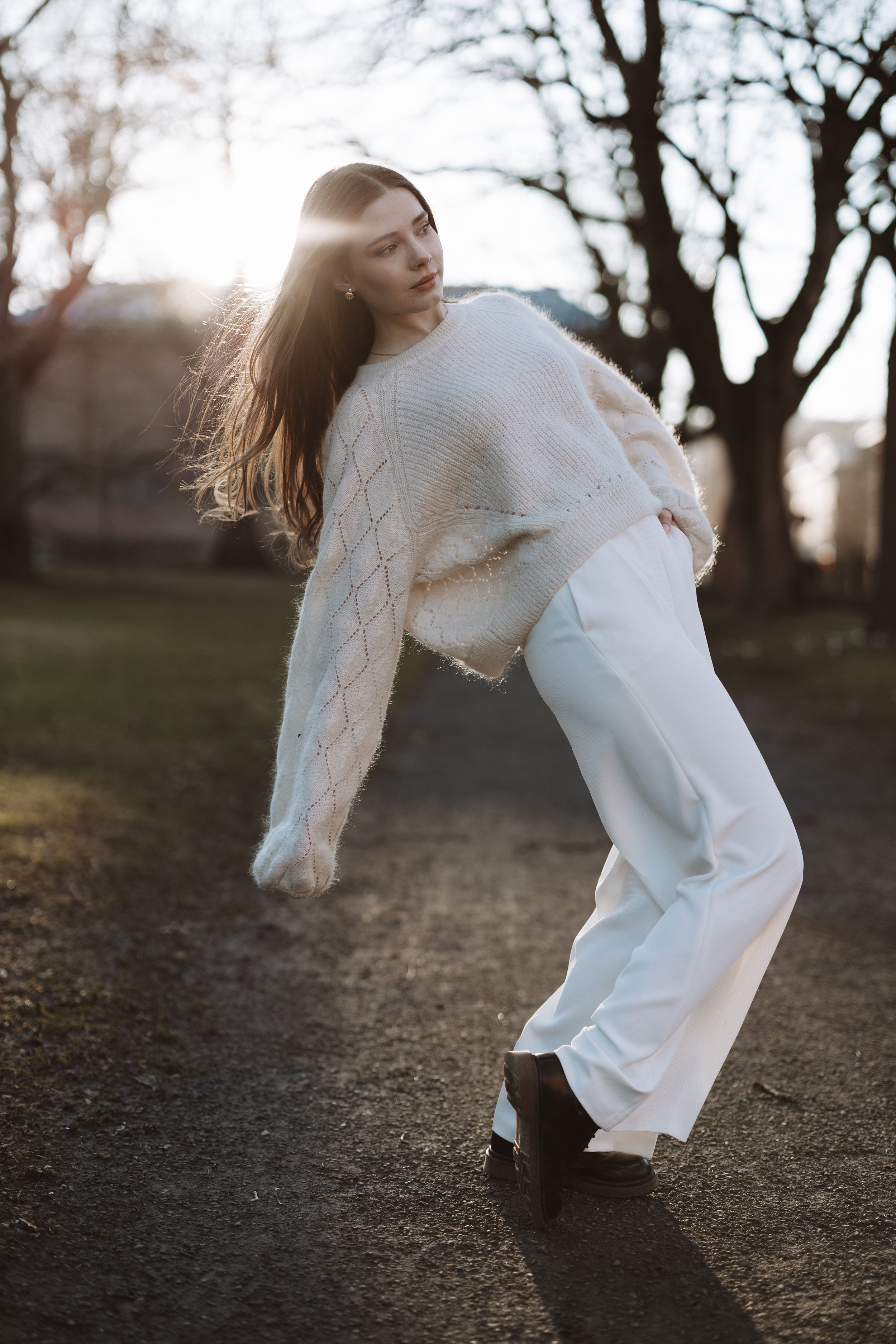 Profesional portrait of woman with long brown hair wearing a white knit sweater and white wide-leg pants, standing in park in Oslo on a dirt path with trees in the background during sunset, striking a stylish pose with sunlight shining behind her.