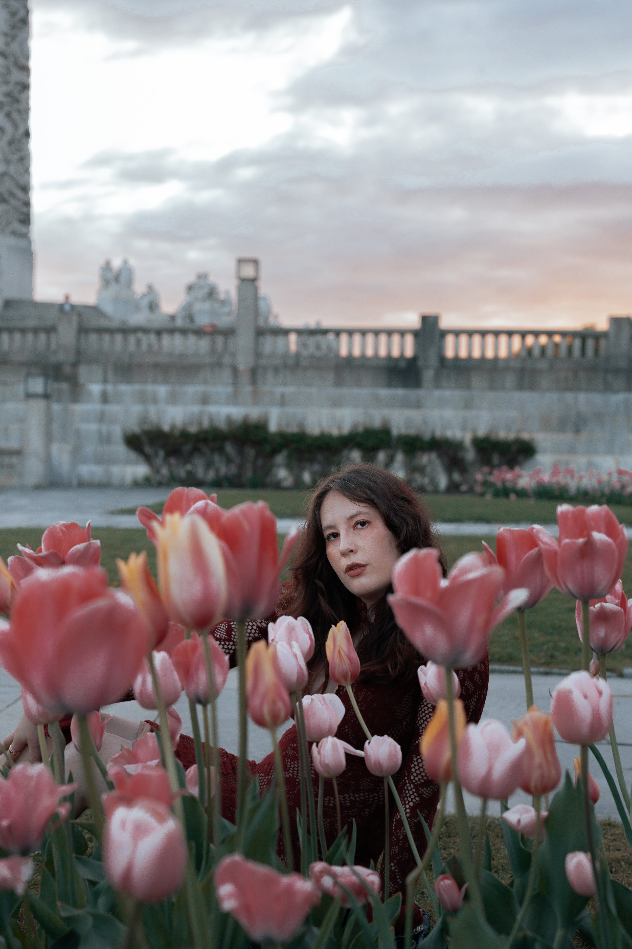 A woman with long dark hair and a maroon dress sits among pink tulips in a garden, with a stone wall and cloudy sky in the background.