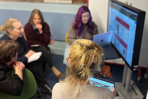 Group of five women engaged in a meeting or presentation, with three women seated on green chairs, one woman with purple hair sitting at a table with a laptop, and another woman with blonde hair using a tablet, in front of a large monitor displaying a presentation.