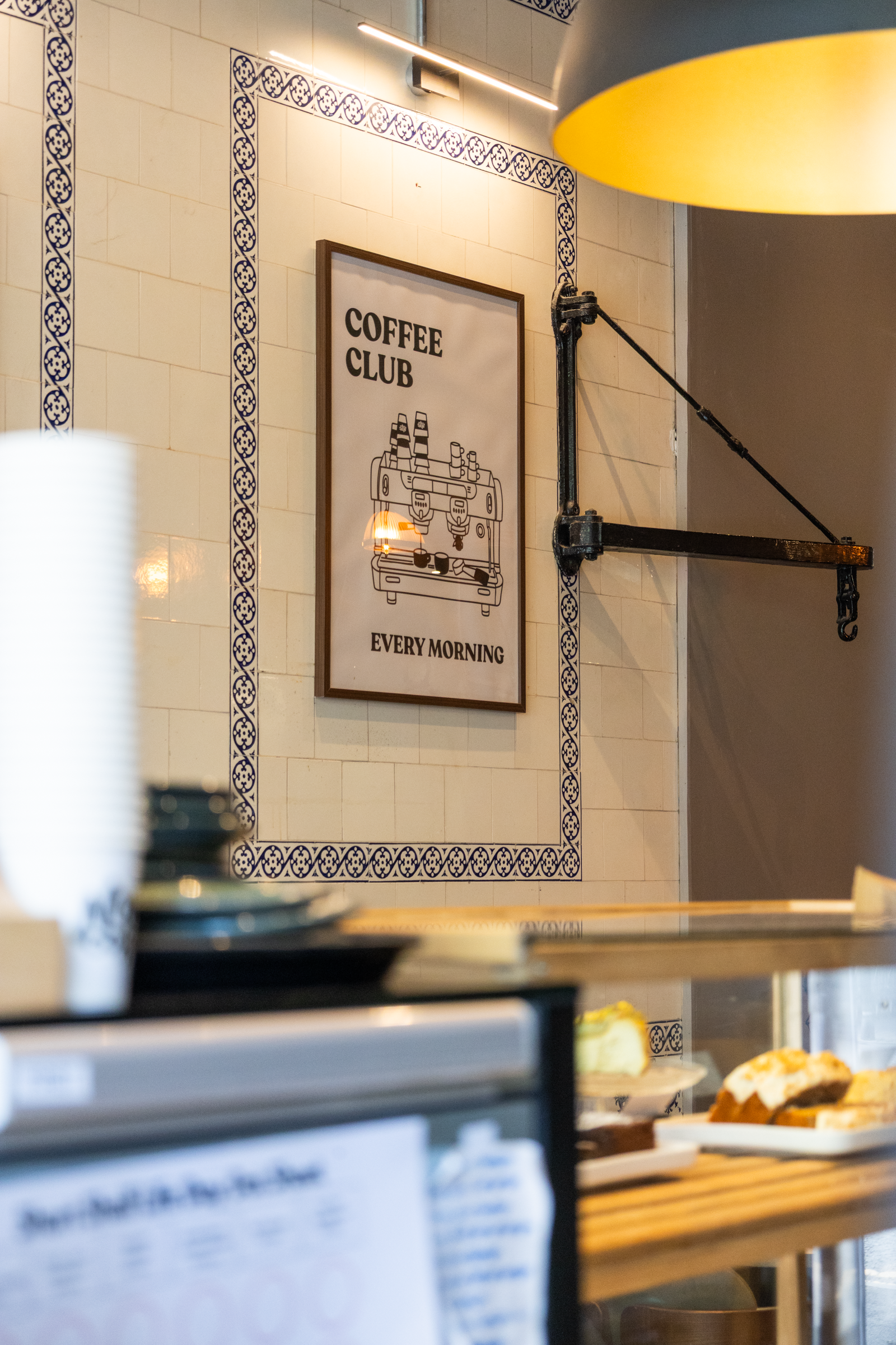 Coffee shop interior with a sign that says "Coffee Club Every Morning" and a coffee machine illustration on the wall. There are some pastries on a table in the foreground.