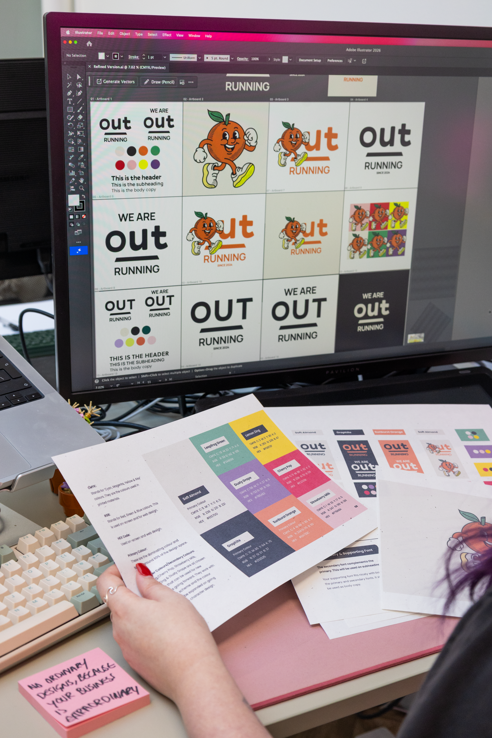 A computer screen displaying various graphic designs for a project featuring a cartoon orange character and the word 'out' in bold font. Several sheets of printed design materials are on the desk, including color palettes and layout samples. A person's hand is holding one sheet of paper.