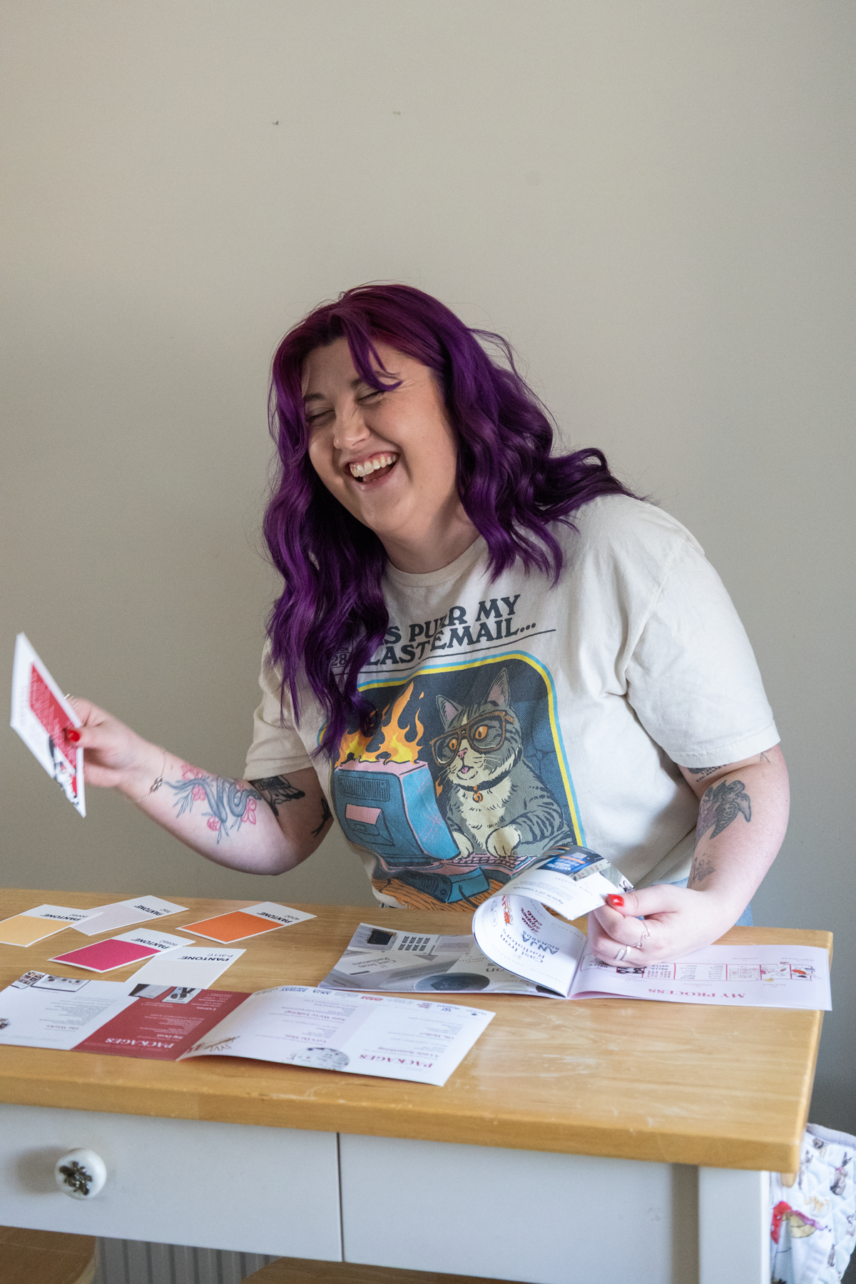 A woman with purple hair laughing and holding papers, sitting at a wooden table with various brochures and color samples, against a plain beige wall.