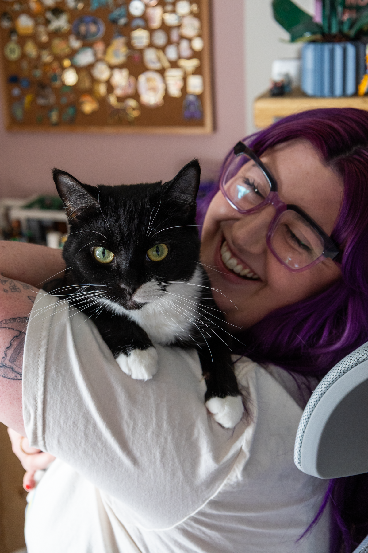 A woman with purple hair and glasses happily holding a black and white cat, with a blurred background of a bulletin board and indoor decor.