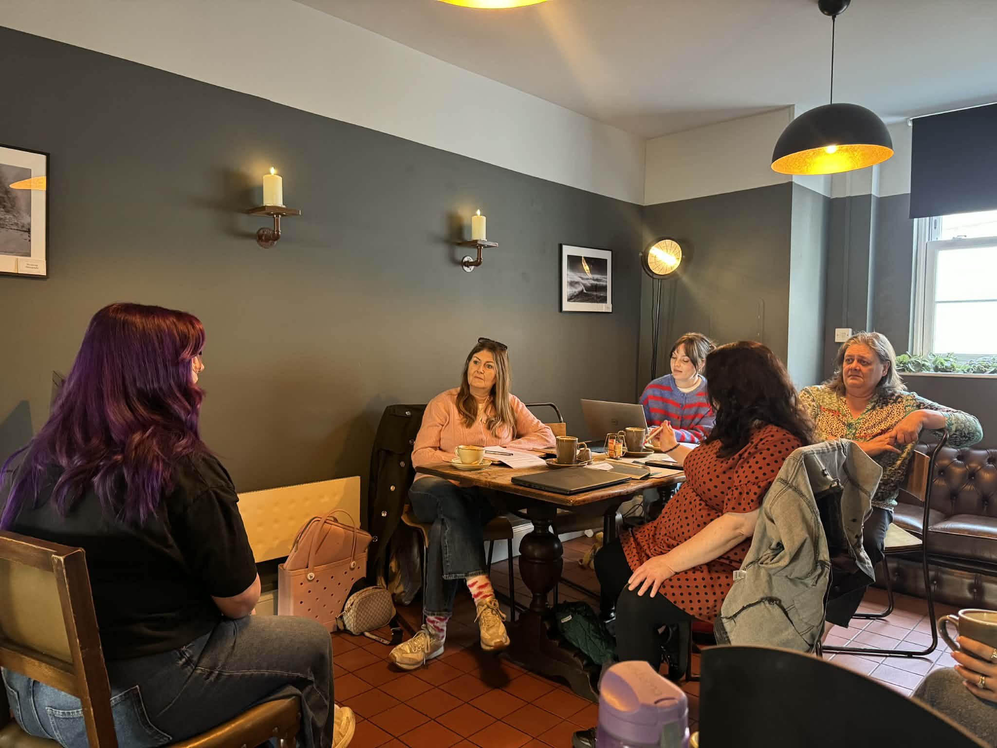 A group of people sitting at a table in a cozy cafe or restaurant, engaging in conversation. The room has dark walls with framed pictures, candles on the walls, and a large hanging light fixture. Windows let in natural light, and the atmosphere is warm and inviting.