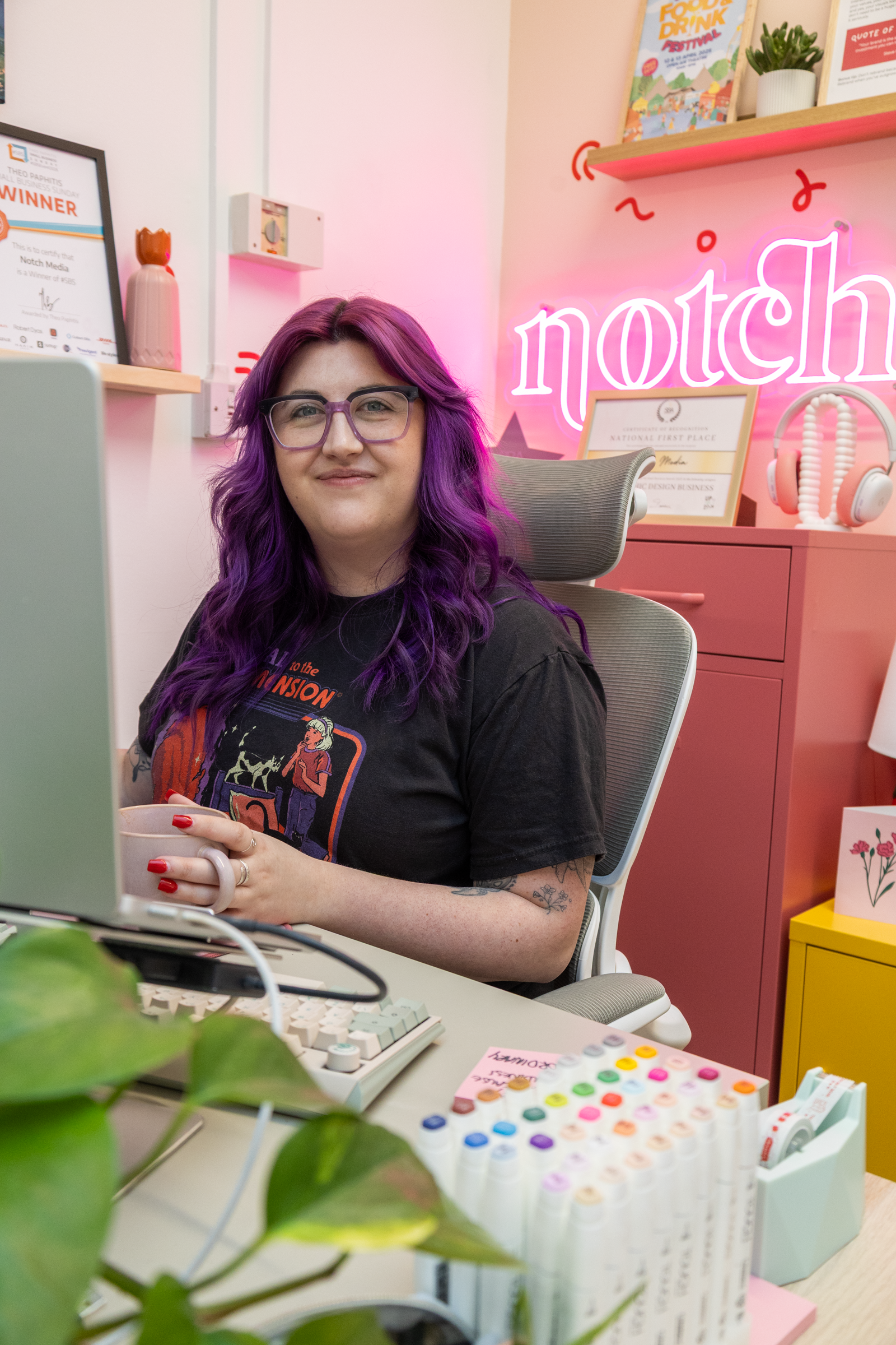 Woman with purple hair and glasses sitting at a desk, smiling, in a colorful, decorated office space with pink neon sign that reads 'notch', and various office supplies and decorations around.