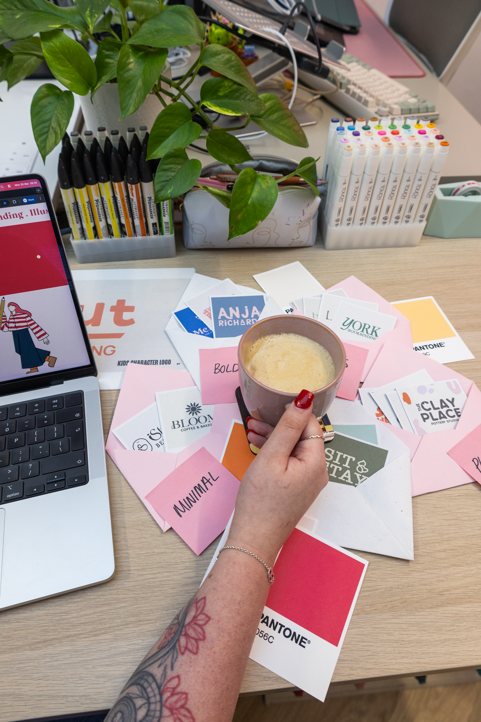 A person's hand holding a coffee mug above a cluttered desk with color swatches, business cards, markers, a laptop, and a plant. The desk displays various office supplies and design samples.