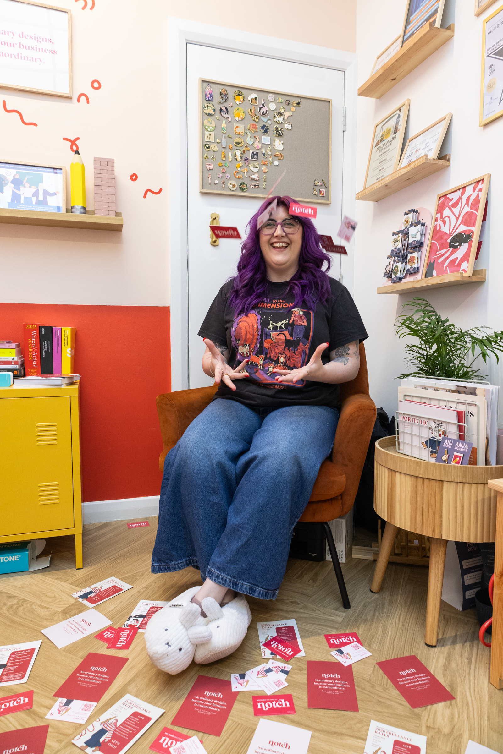 A woman with purple hair and glasses is sitting on an orange chair, smiling, in a room decorated with framed art and shelves. She is wearing a black graphic T-shirt and blue jeans. White plush slippers shaped like a bunny are on her feet. The floor around her has scattered red and white brochures and cards, some on the table nearby.