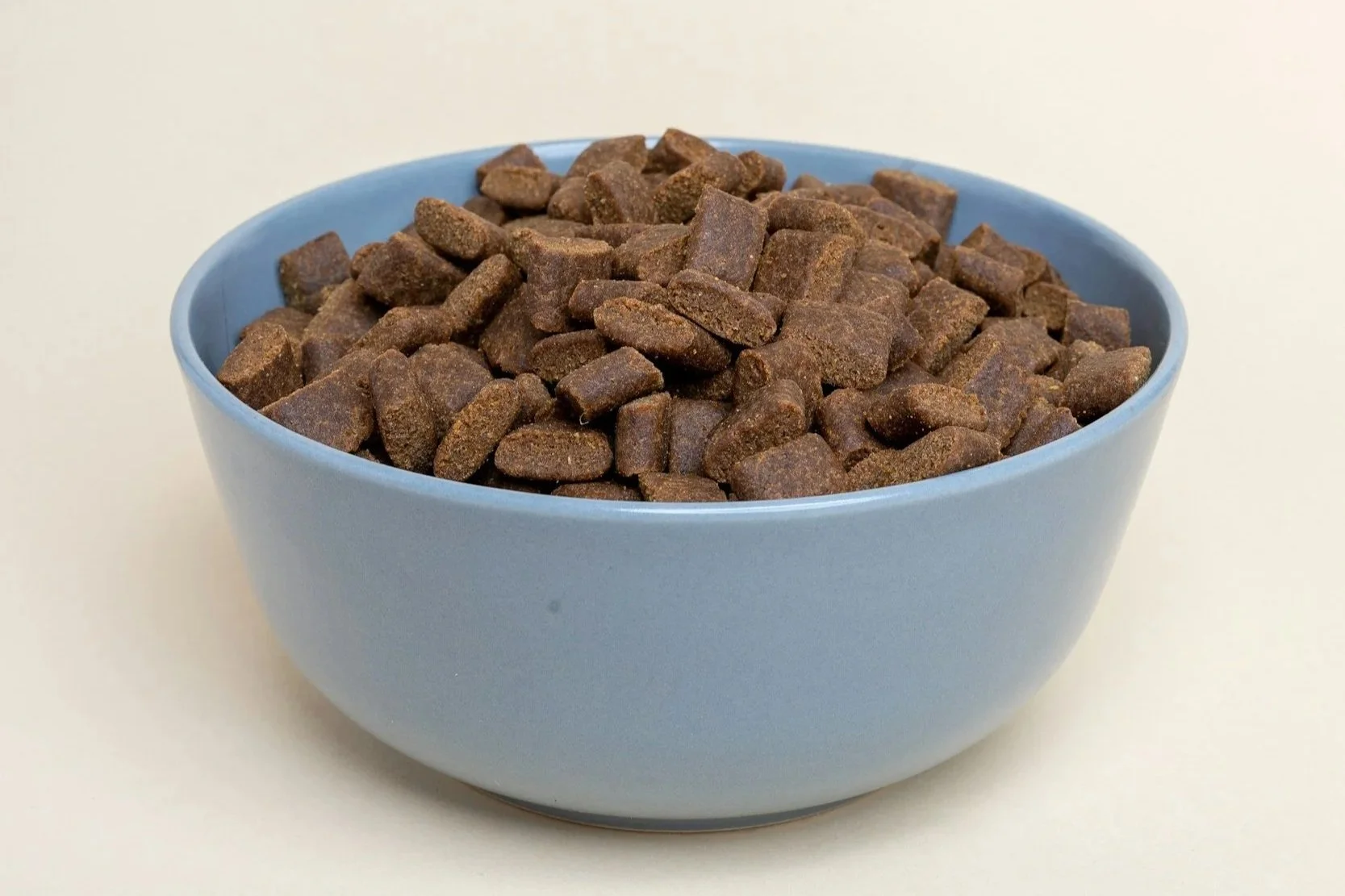 Blue bowl filled with small, brown, rectangular dog treats on a plain white background.