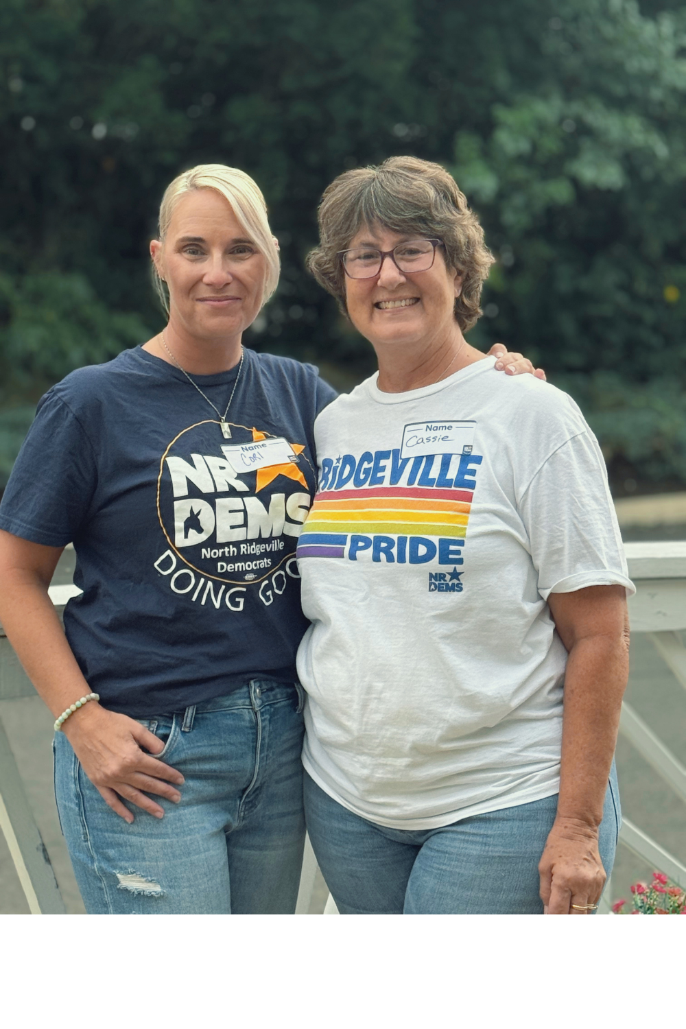 Two women standing outdoors on a bridge, smiling, with green trees in the background. Both are wearing political shirts supporting the North Ridgeville Democrats. The woman on the left has blonde hair, a navy blue shirt, and a name tag that says Carl. The woman on the right has short, gray hair, wears glasses, and a white shirt with a rainbow-colored logo that says "BRIDGEVILLE PRIDE," and a name tag that says Cassie.