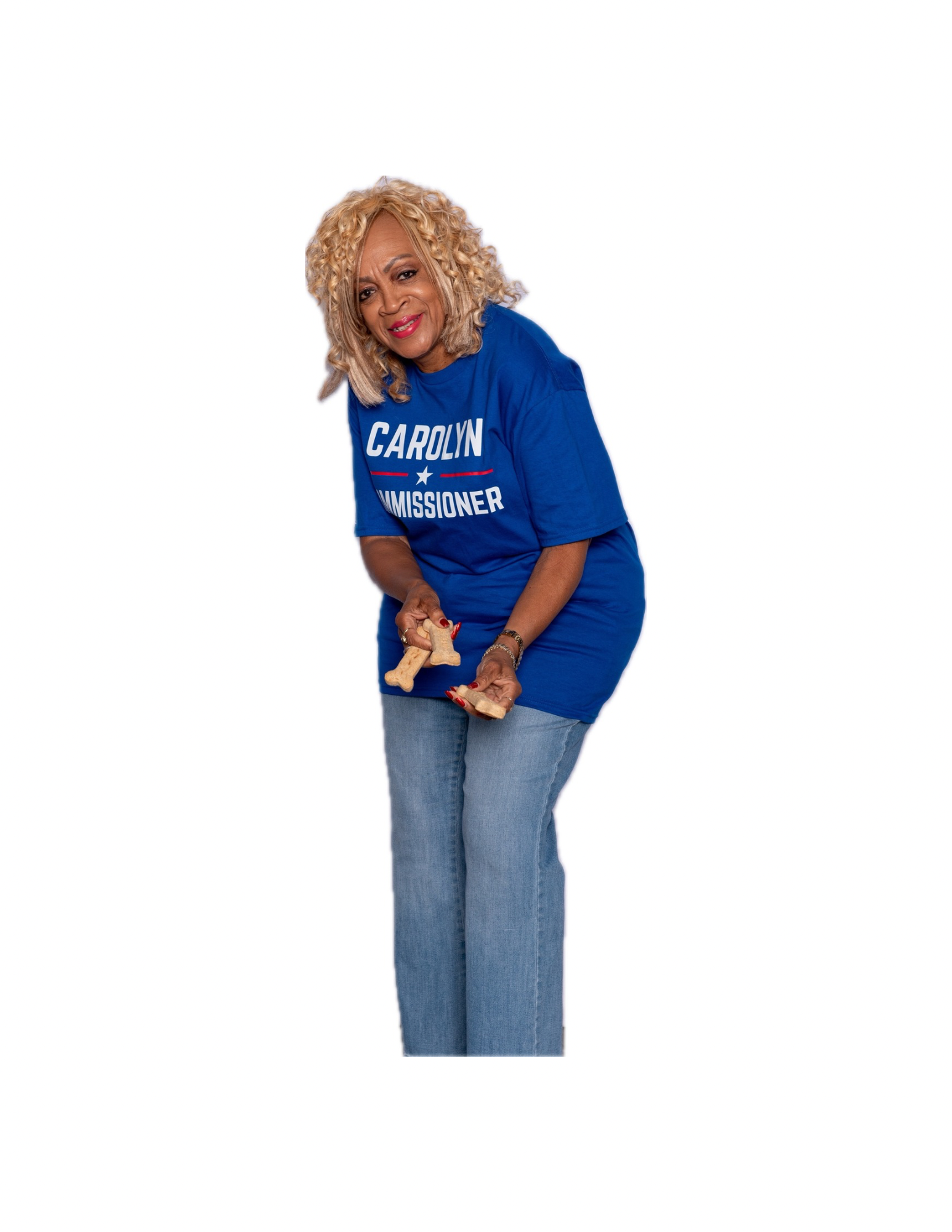 A woman with blonde curly hair smiling, wearing a blue campaign t-shirt that reads 'Carolyn Commissioner,' and holding a rolled-up item in her hands, standing against a white background.