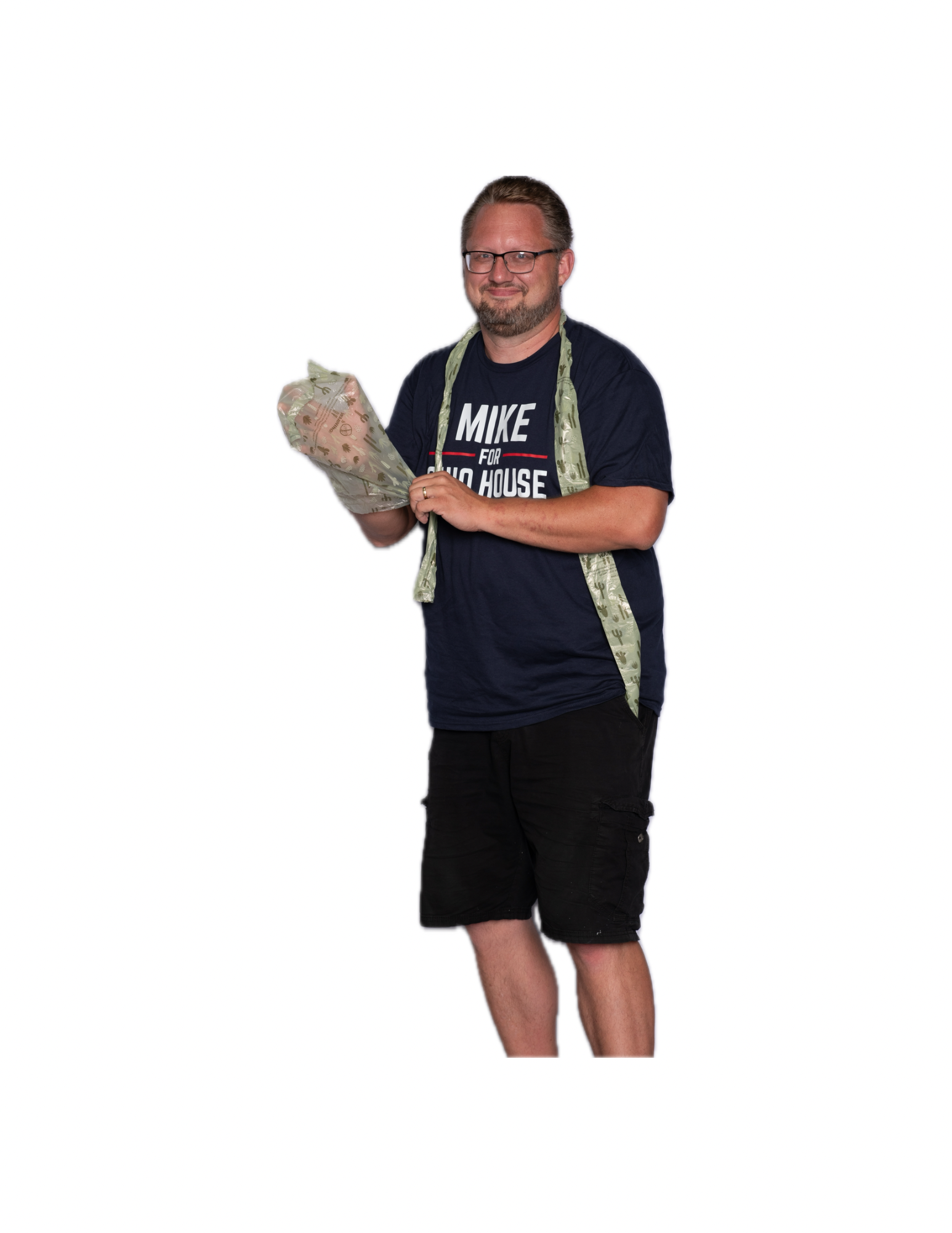 Man with glasses, beard, and mustache wearing a dark blue t-shirt with text, shorts, and a green patterned apron, holding a wrapped giftbox, standing against a white background.