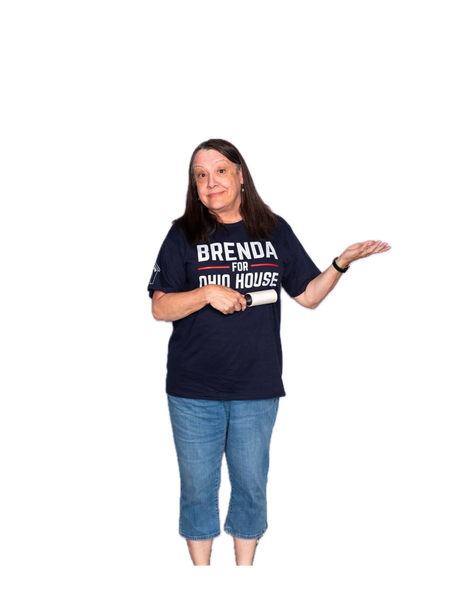 A woman with long dark hair and a light skin tone, wearing a navy blue T-shirt with political campaign text, jeans, and holding a small black object in her right hand, standing against a plain white background.