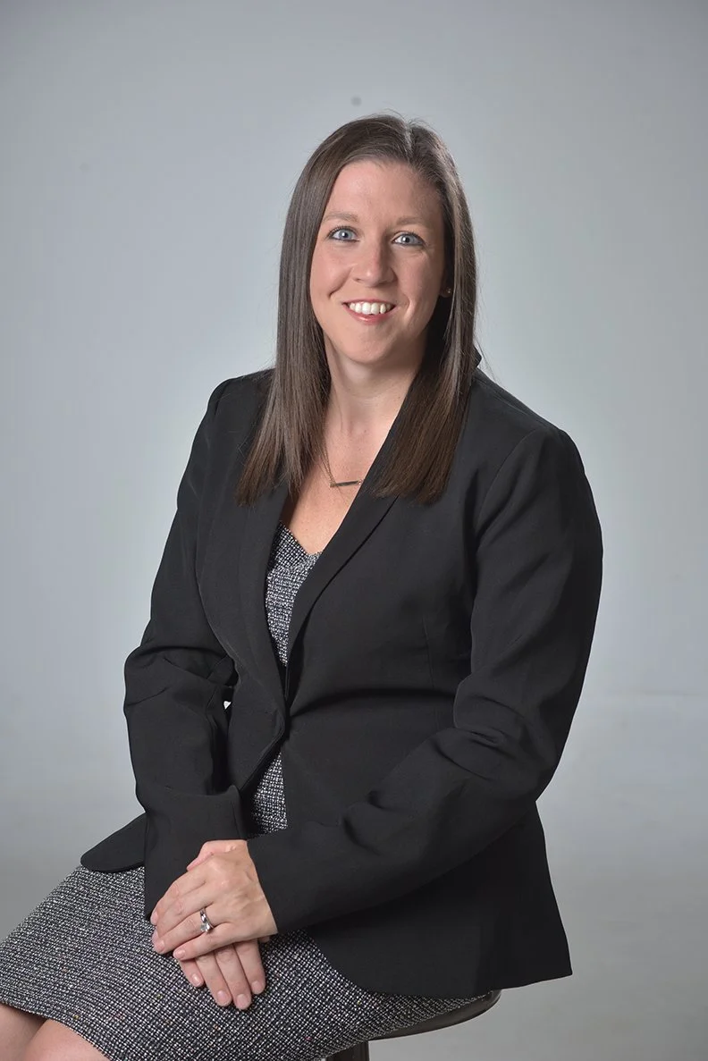 A professional woman with shoulder-length brown hair and blue eyes, wearing a black blazer and a gray patterned dress, sitting on a stool against a plain gray background with a slight smile.