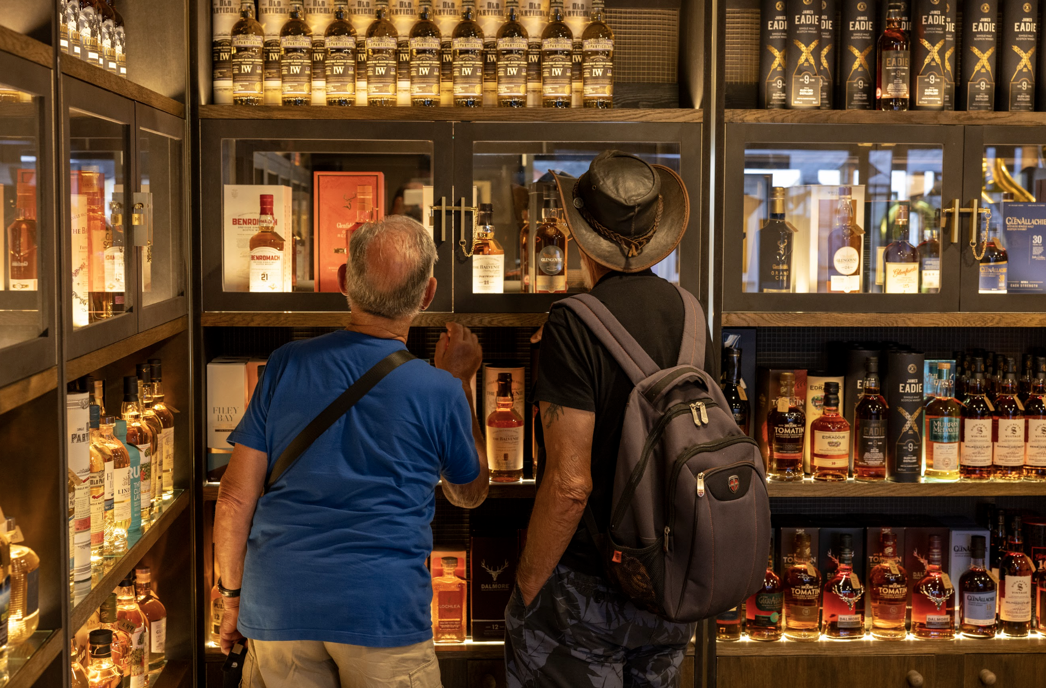 Two men shopping for whiskey in a liquor store, with shelves full of whiskey bottles behind them.